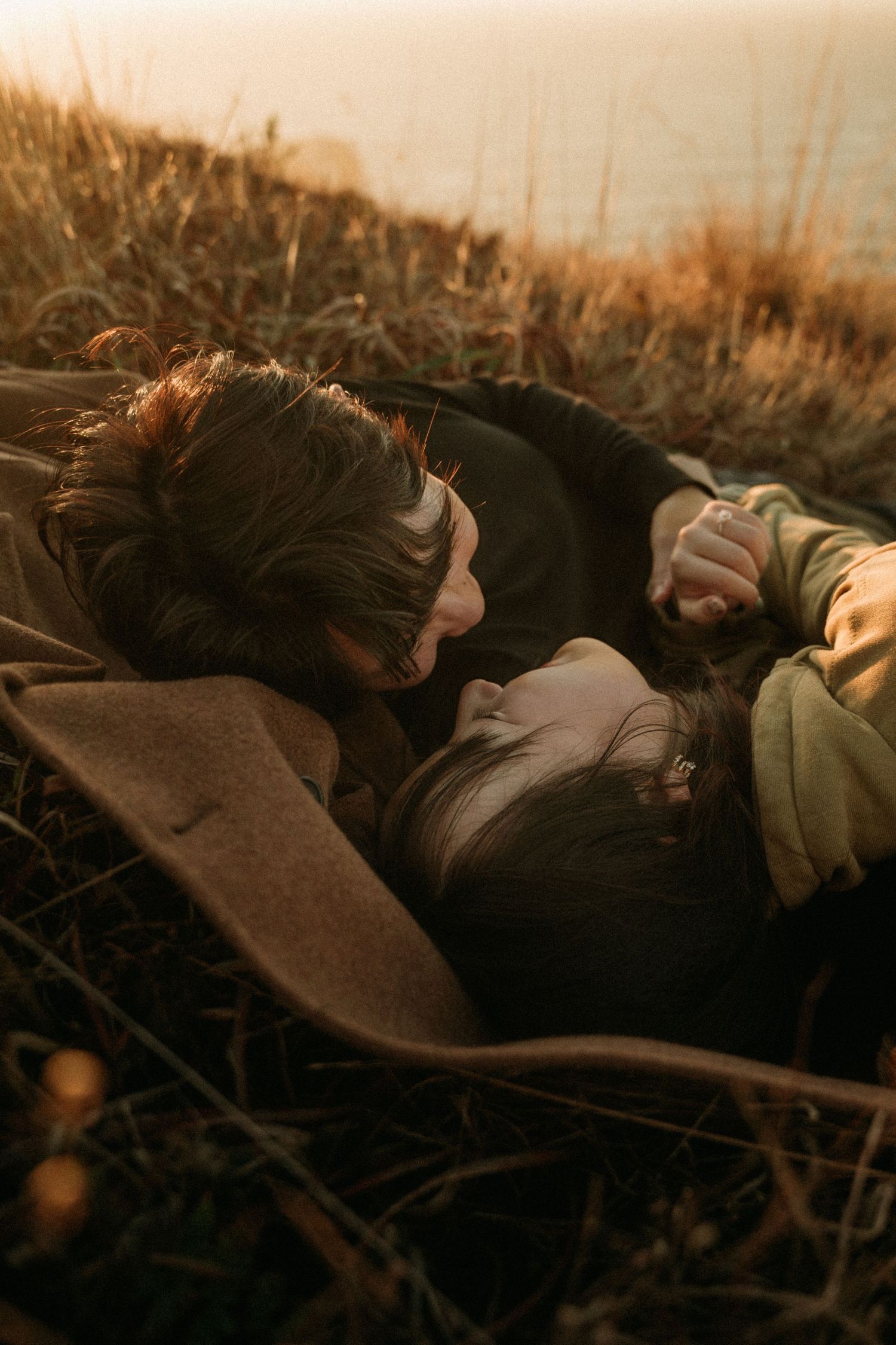 Couple lying close together on a grassy hillside at golden hour, wrapped in a tan coat, sharing a tender moment.