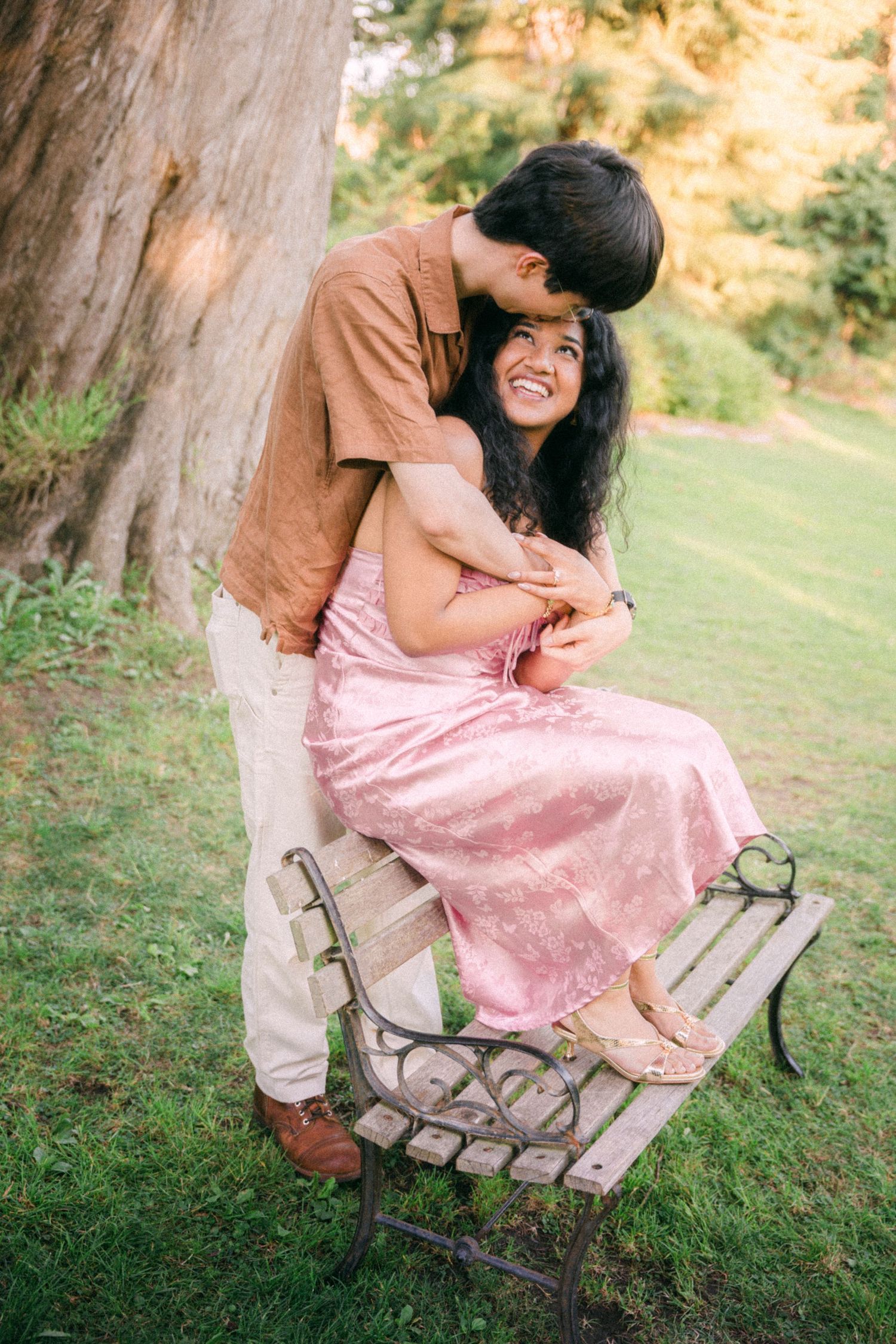 Couple sharing a romantic moment on a park bench, woman in pink dress smiling as man kisses her forehead.