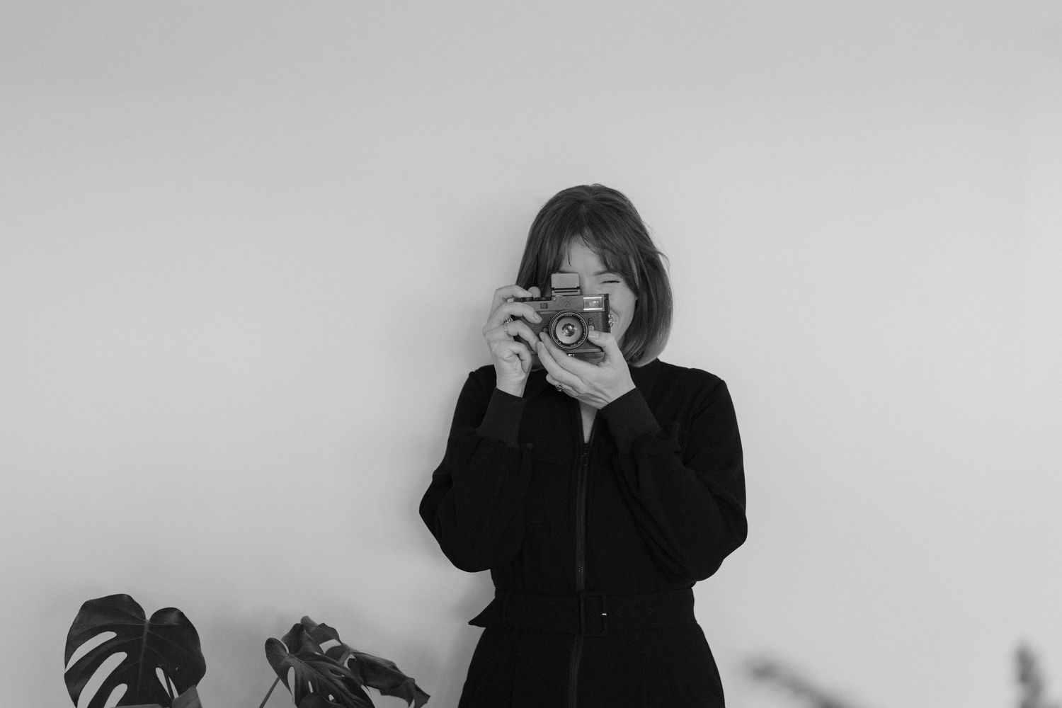 Black and white photo of a woman in black holding a vintage camera up to her face, with a plant in the background.Photography by The Y.K. St