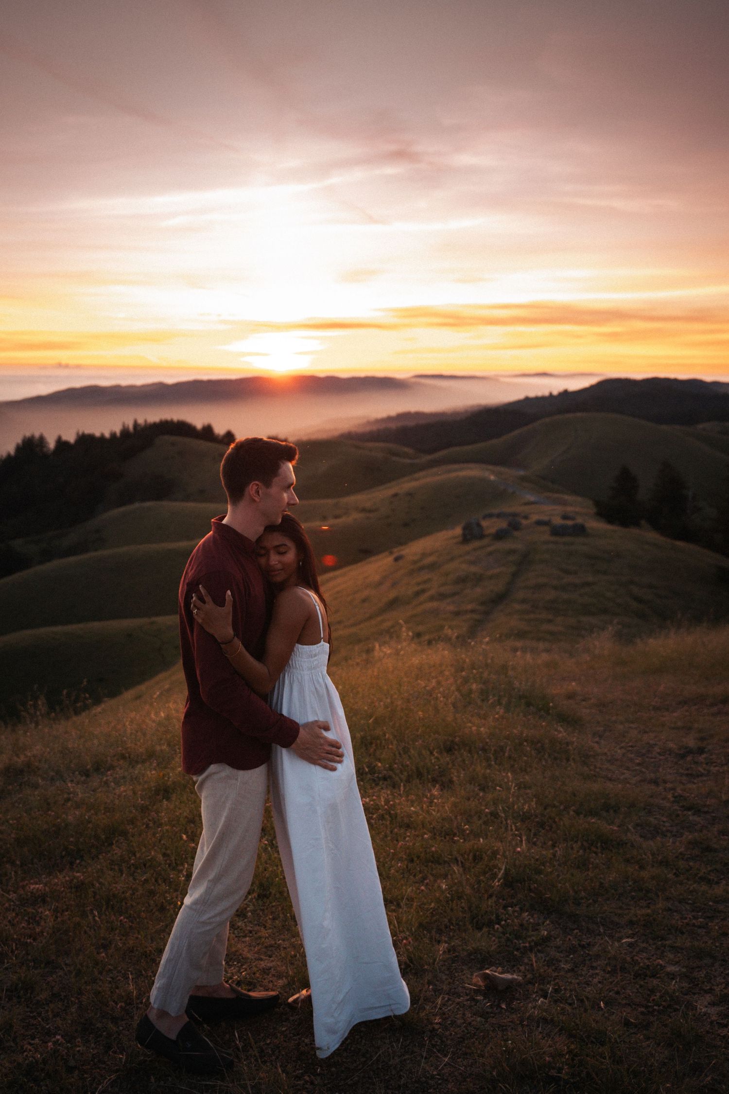 Couple embracing at golden sunset on green hills, woman in white dress, man in burgundy shirt, romantic outdoor portrait.Photography by The