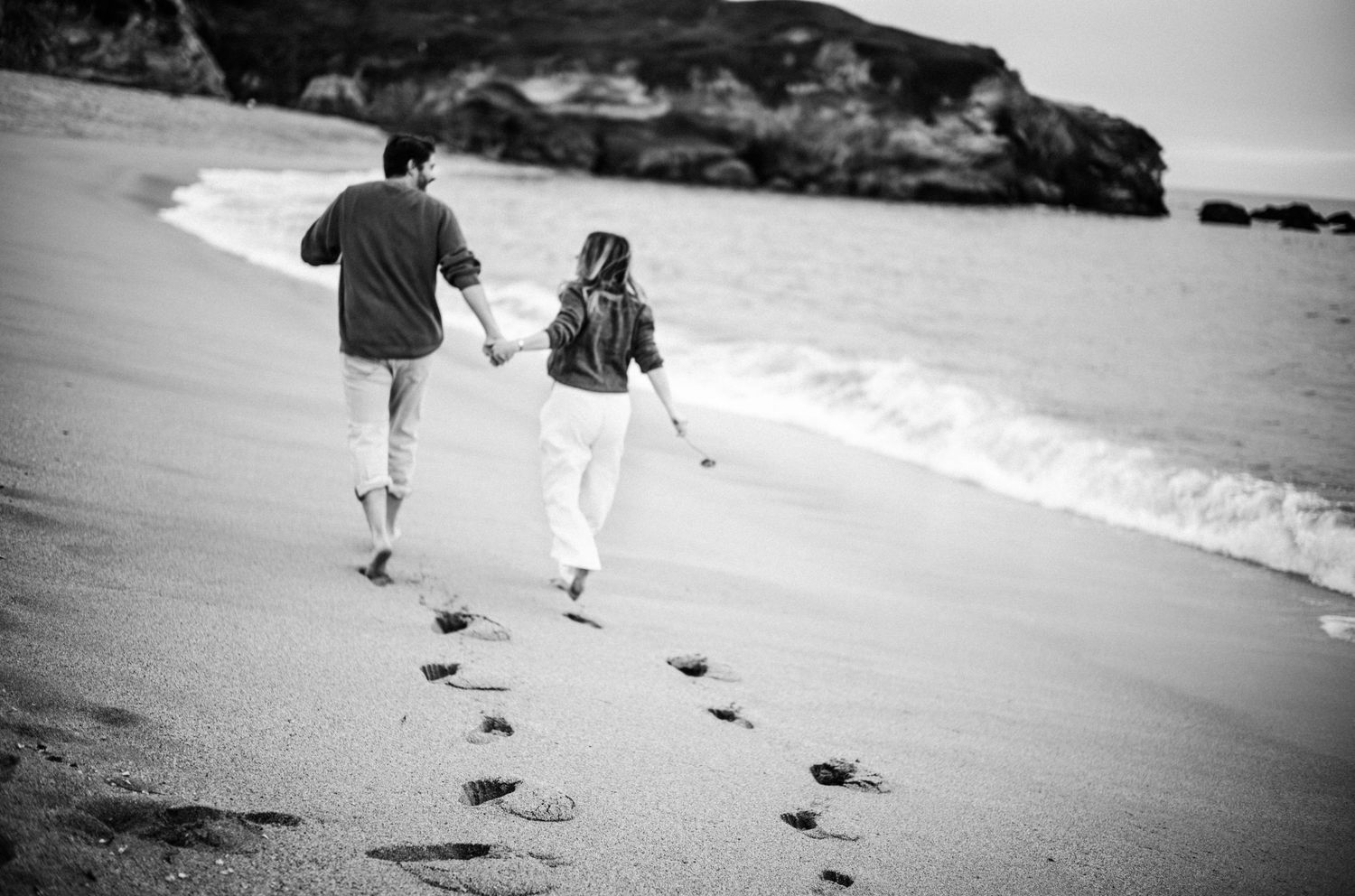 Black and white photo of a couple holding hands and walking along a sandy beach, leaving footprints in the sand.Photography by The Y.K. Stud