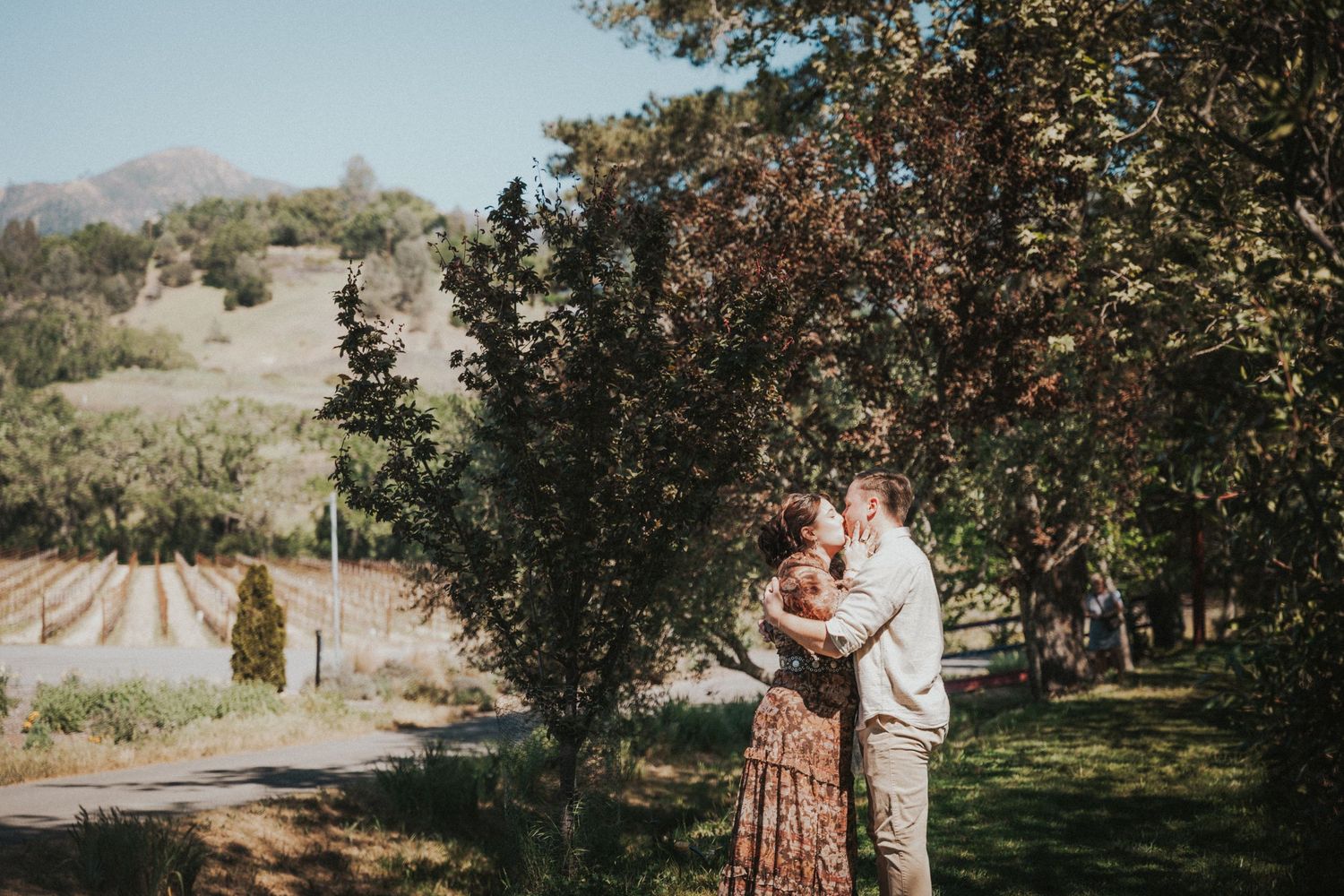 Couple embracing romantically among lush trees at a scenic vineyard on a sunny day.