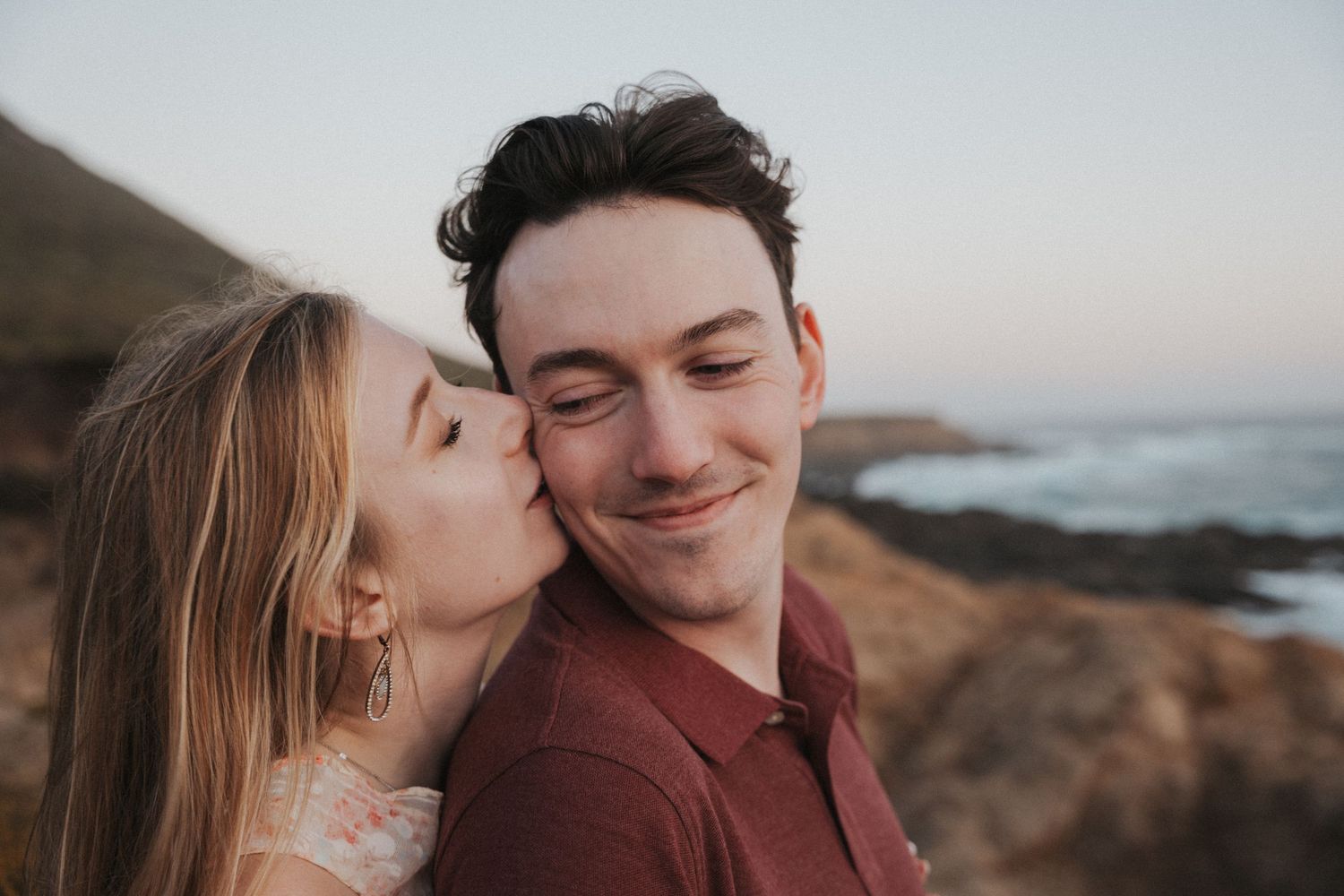 A woman kisses a smiling man on the cheek outdoors near a scenic coastal cliff and ocean backdrop.