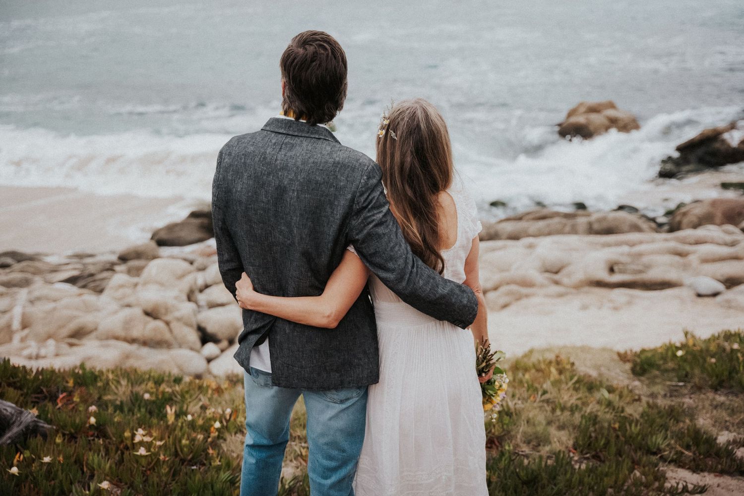 Couple embracing while gazing at rocky ocean shoreline, viewed from behind, wearing casual and white dress.