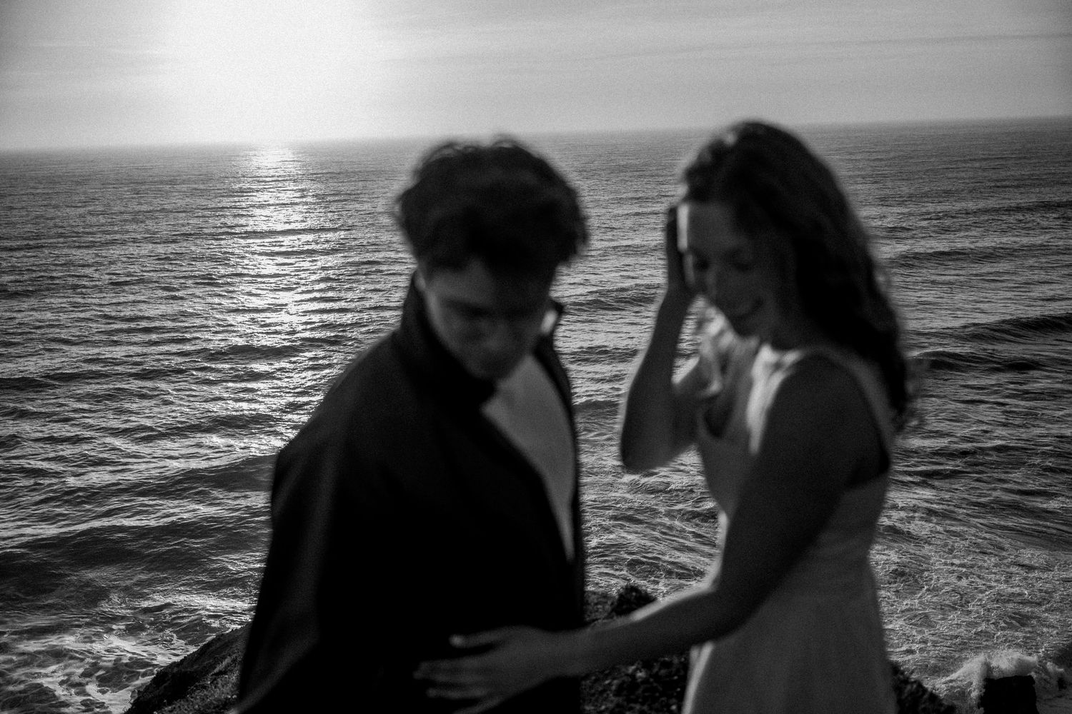 Couple standing on rocky shore at sunset in black and white, woman touching hair by the ocean.