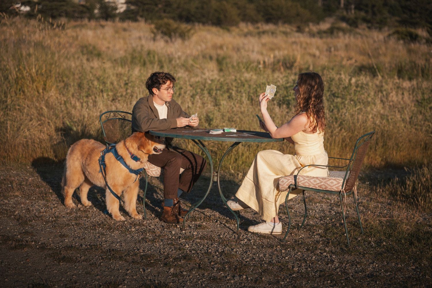 Couple sitting at outdoor table with golden retriever dog in a sunlit field at golden hour.