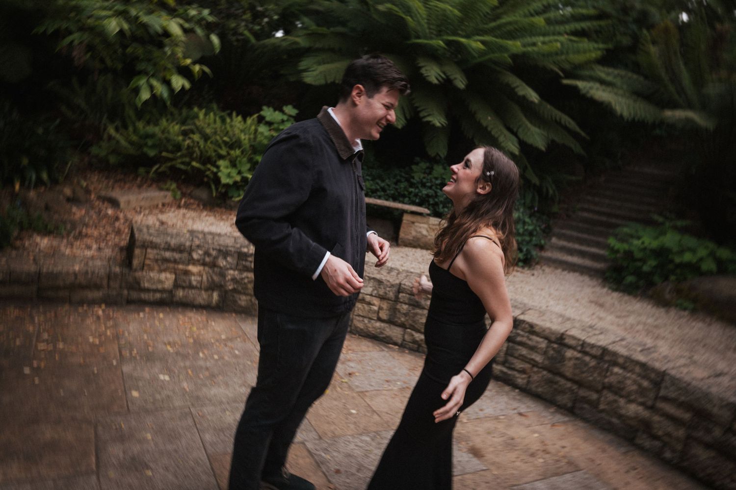 Couple in elegant black attire sharing a romantic moment in a lush garden setting.