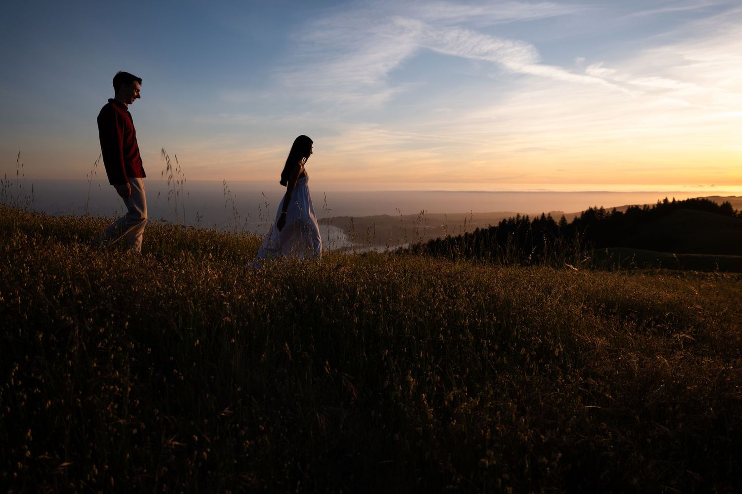 Couple silhouetted against a stunning sunset sky walking on a grassy hilltop with mountain views.