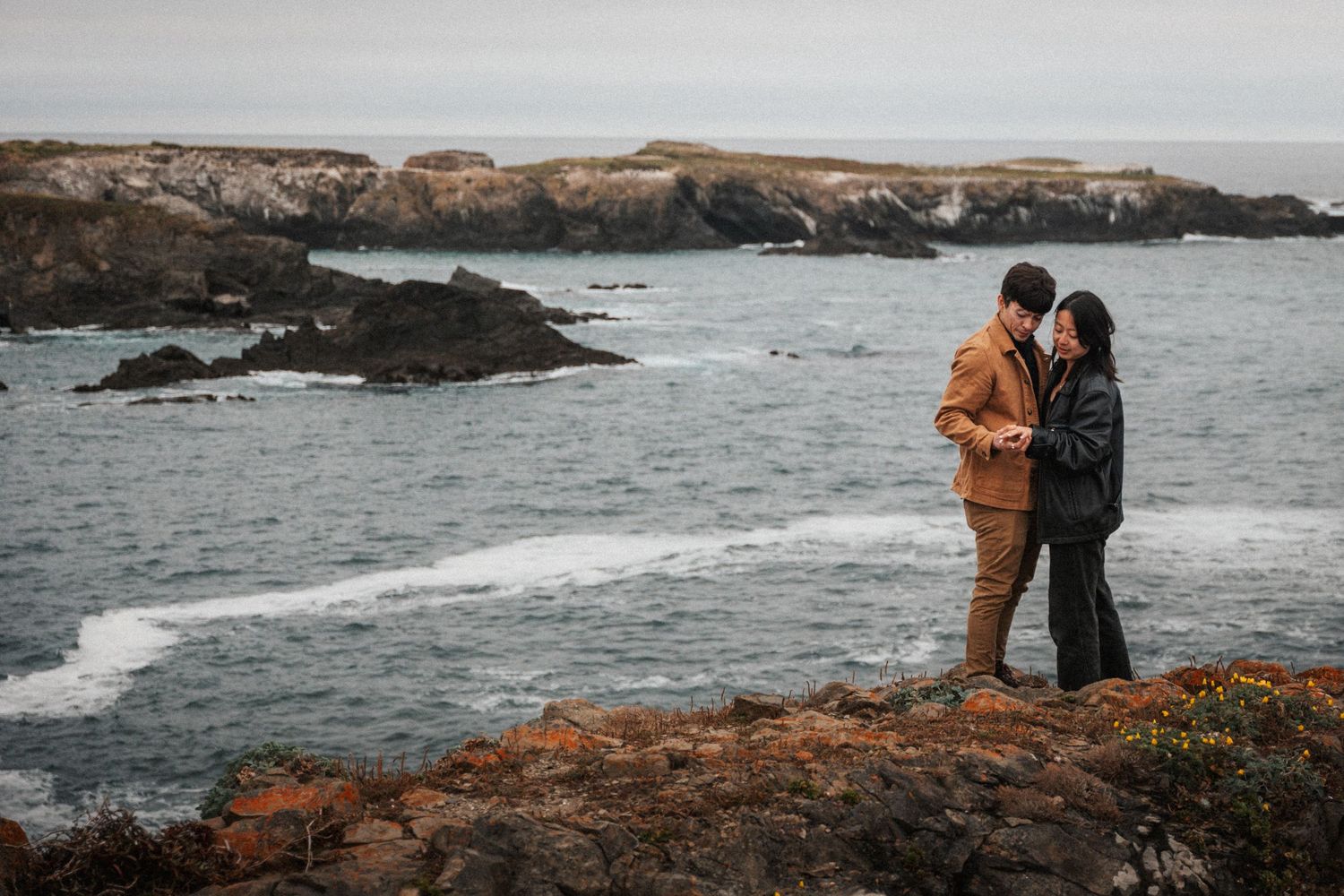 Romantic couple standing on rocky coastal cliffs overlooking dramatic ocean waves and sea stacks.
