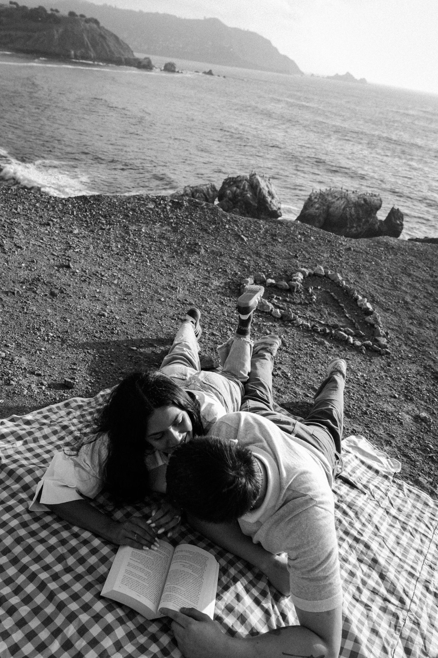 Black and white aerial view of a couple lying on a blanket reading a book on a rocky beach.