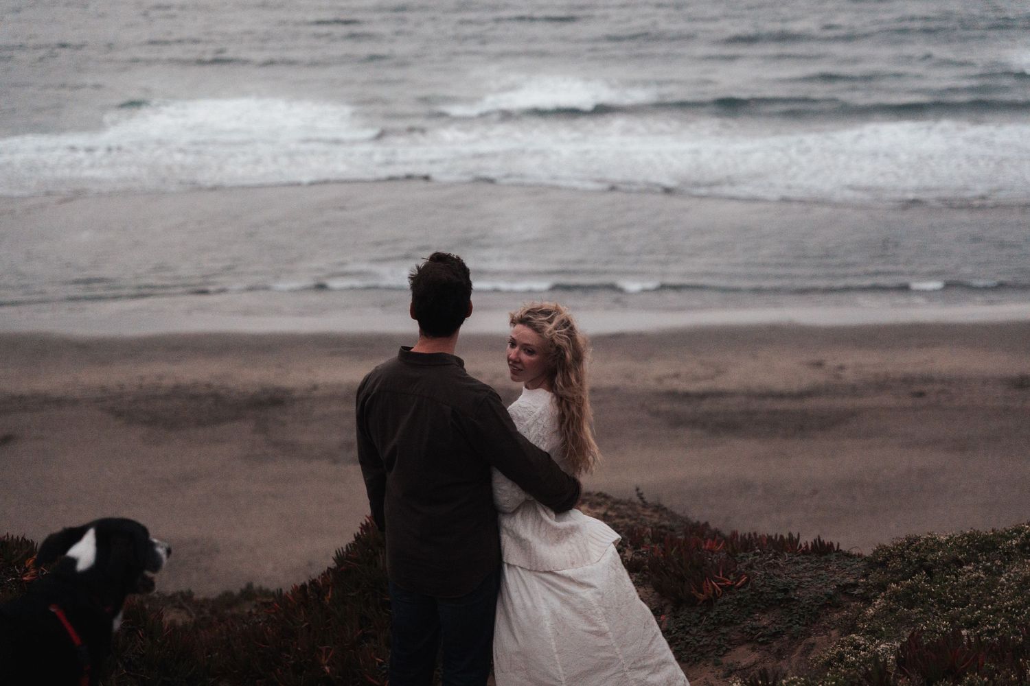 Couple in wedding attire embracing on a beach with ocean waves and a dog nearby.