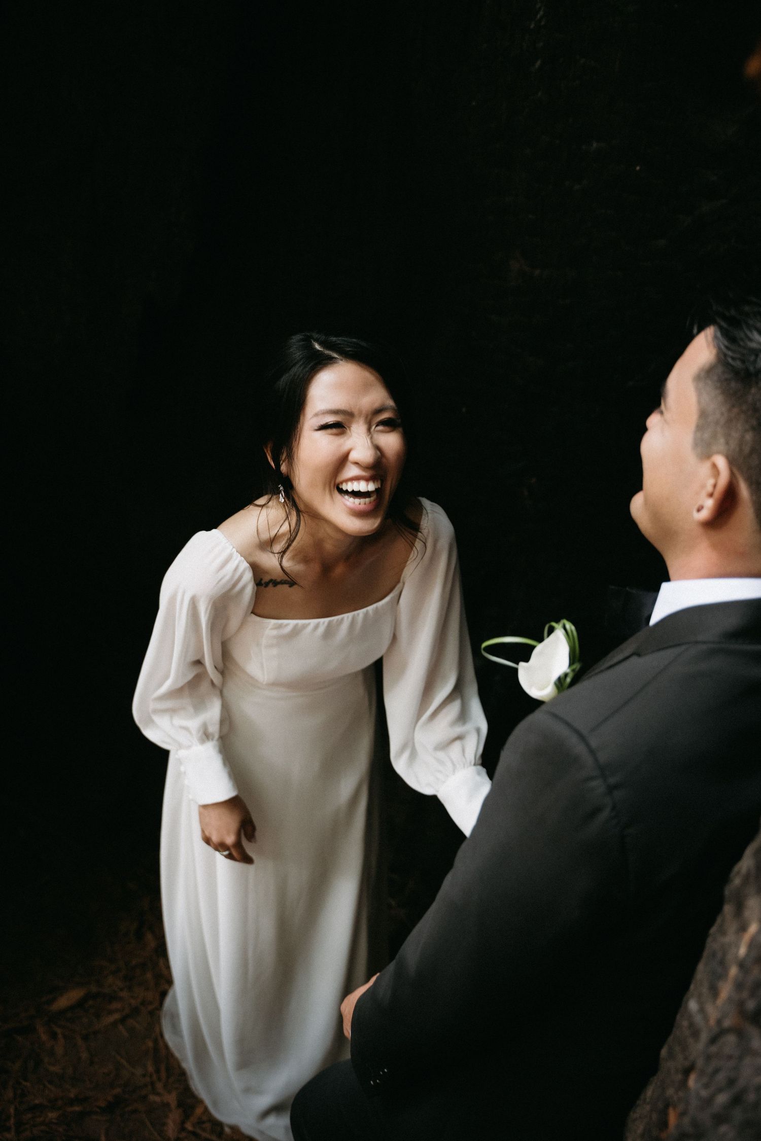Bride in white dress laughs joyfully with groom in black suit during outdoor wedding ceremony.