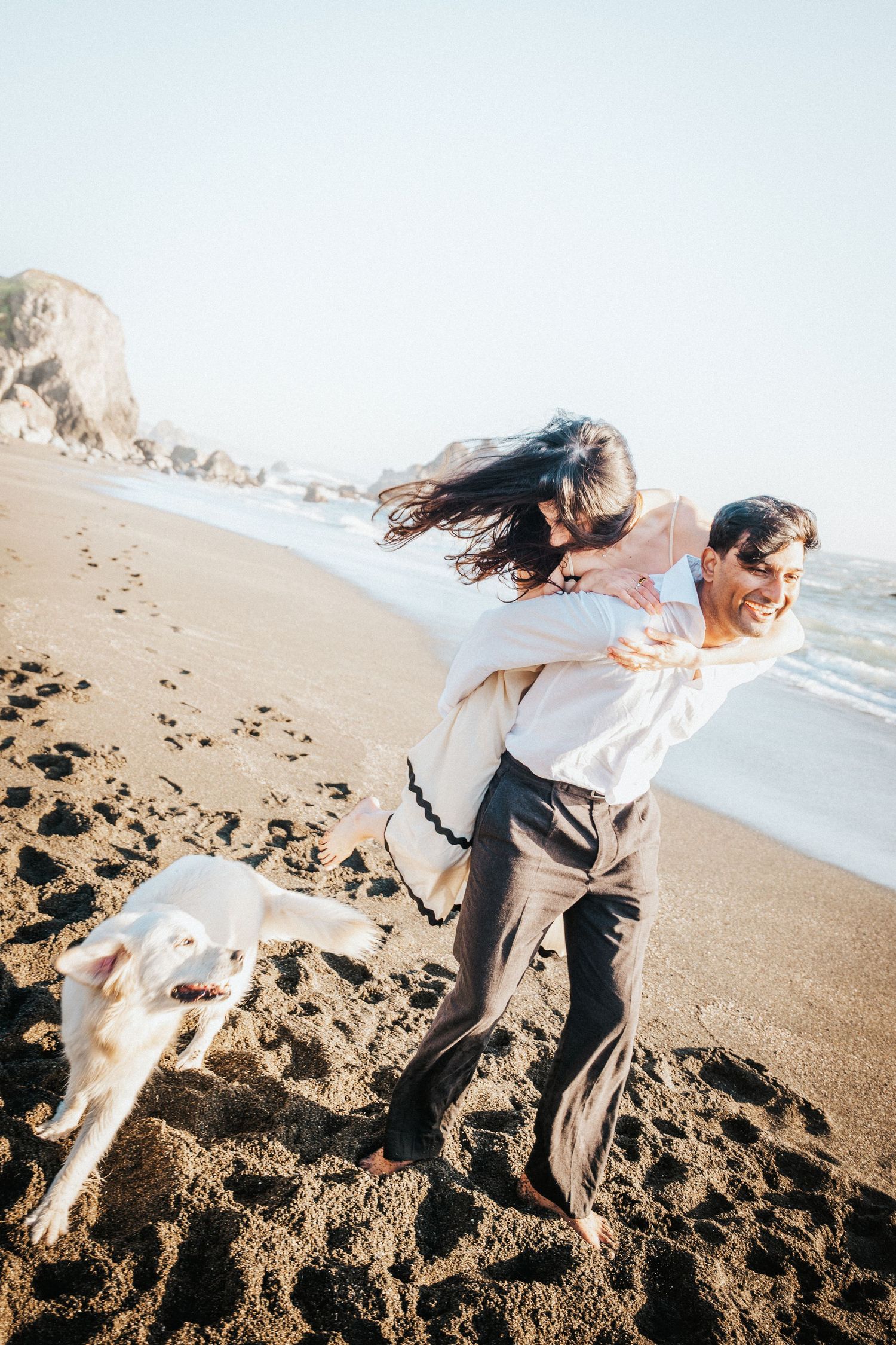 Man giving woman a piggyback ride on a beach while their white dog runs alongside them.