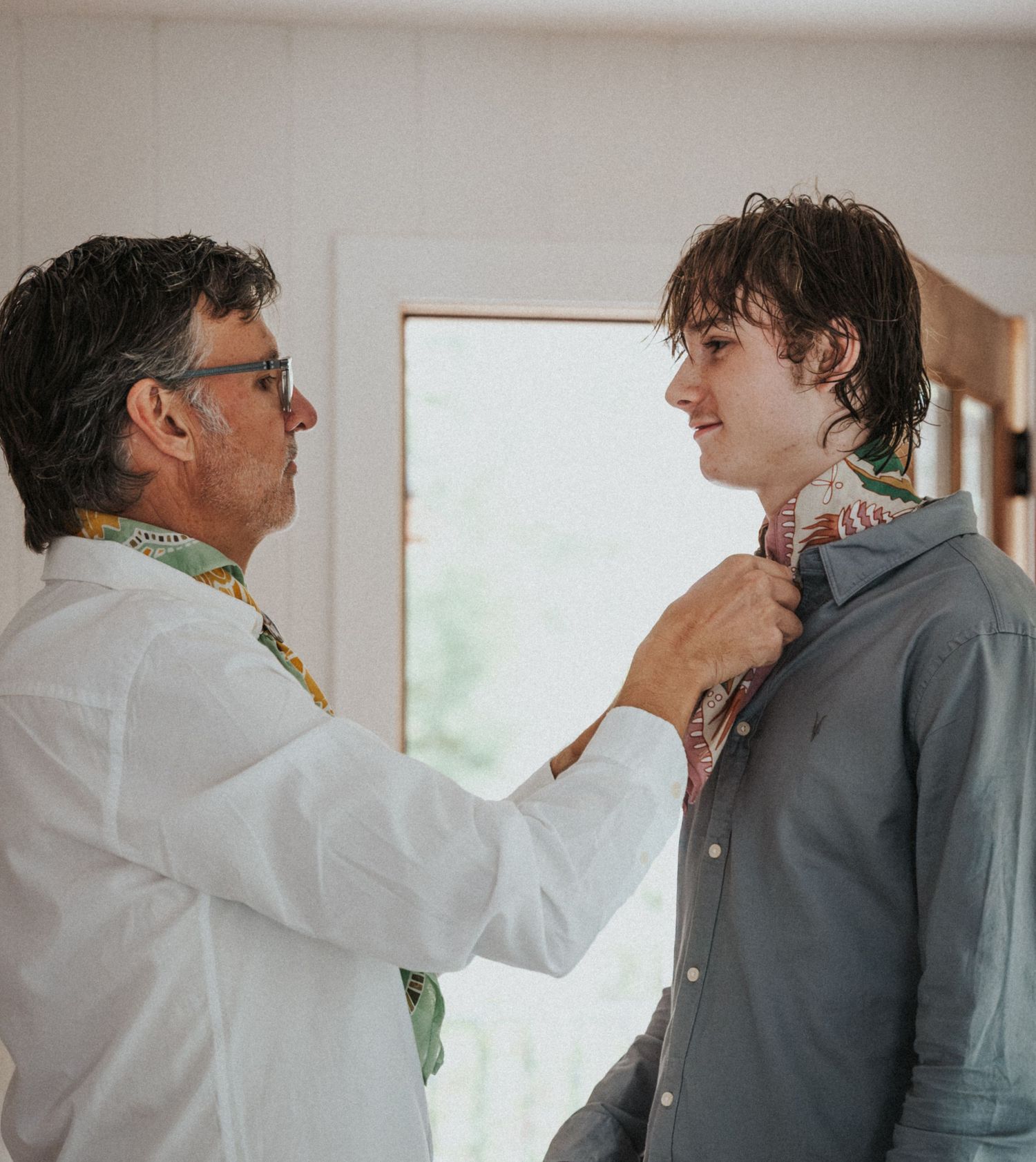 An older man helps a young man adjust his tie before a wedding ceremony indoors.