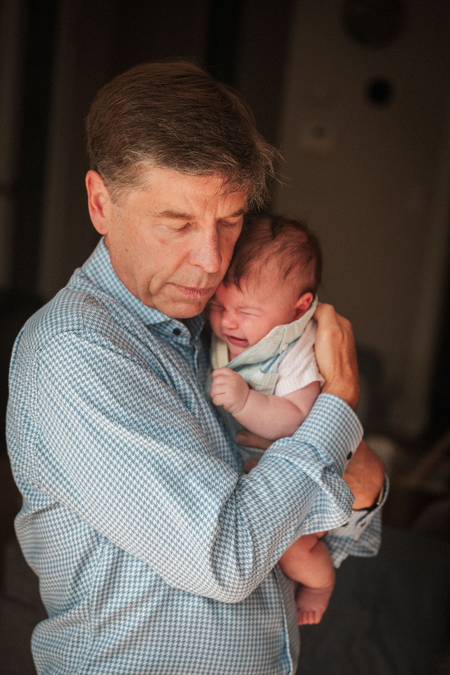 Elderly man in blue plaid shirt tenderly holding a crying newborn baby close to his cheek.
