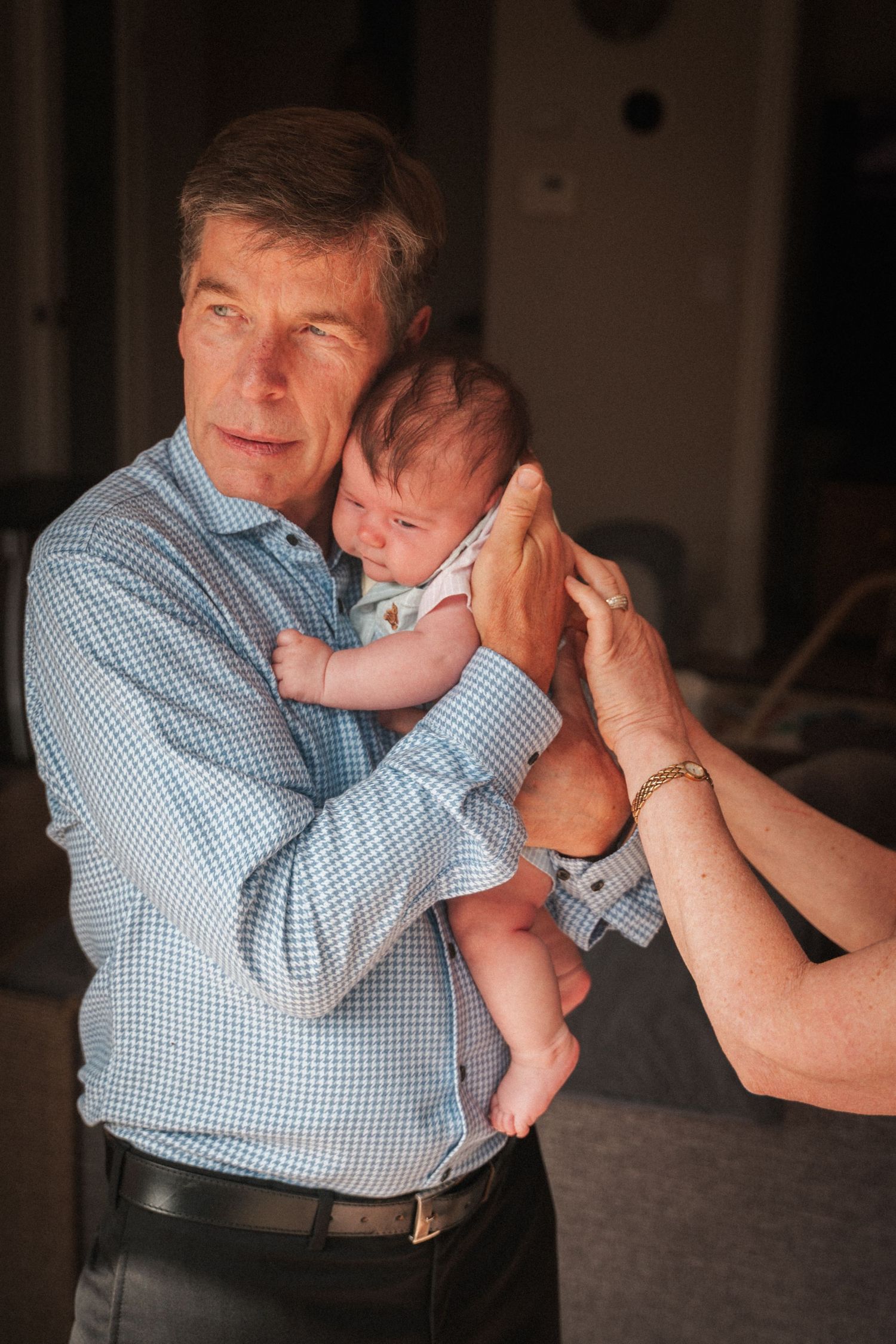 An elderly man tenderly holds a newborn baby while another person gently supports the infant.