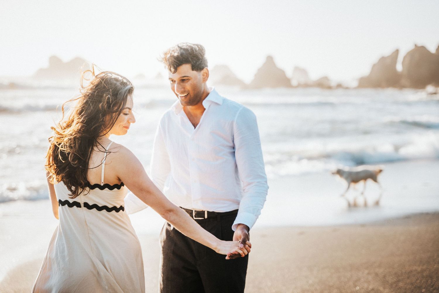 A happy couple holds hands and walks along a sunny beach with waves and a dog in the background.