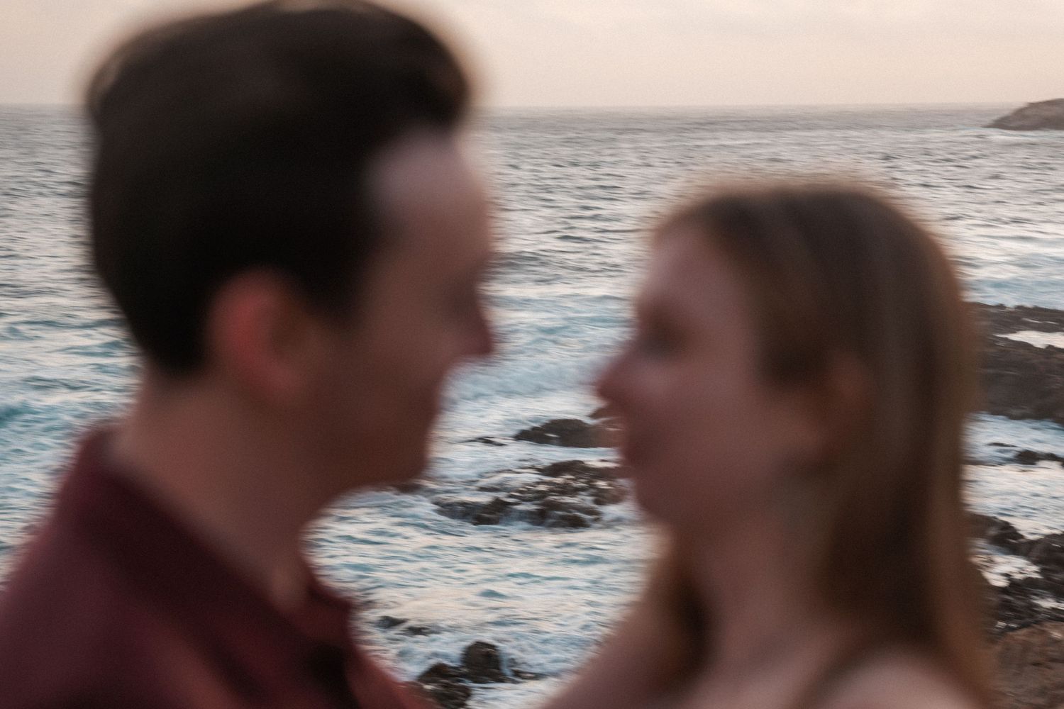 Couple sharing a romantic moment by the ocean at dusk, with rocky coastline and blue water in the background.