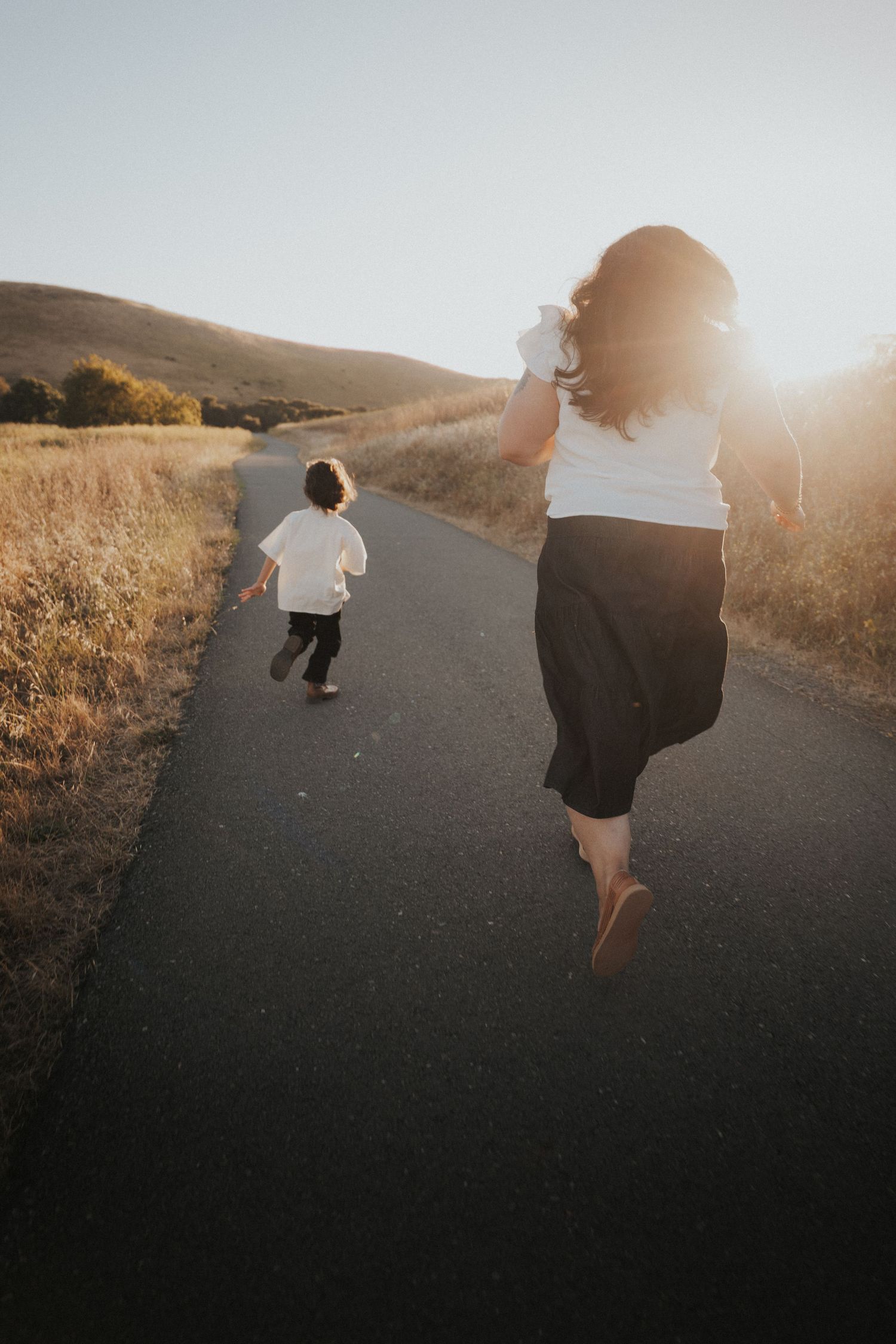 Mother and child running together on a sunlit path through golden fields at sunset.