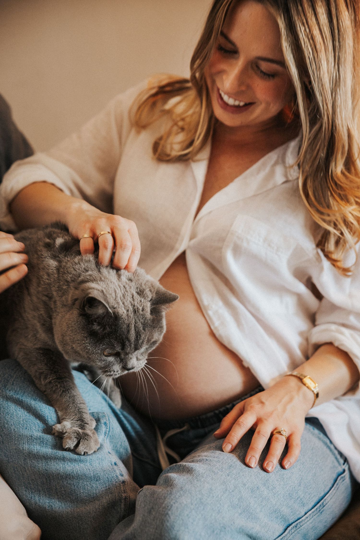Smiling pregnant woman in white shirt petting a gray British Shorthair cat while sitting comfortably at home.