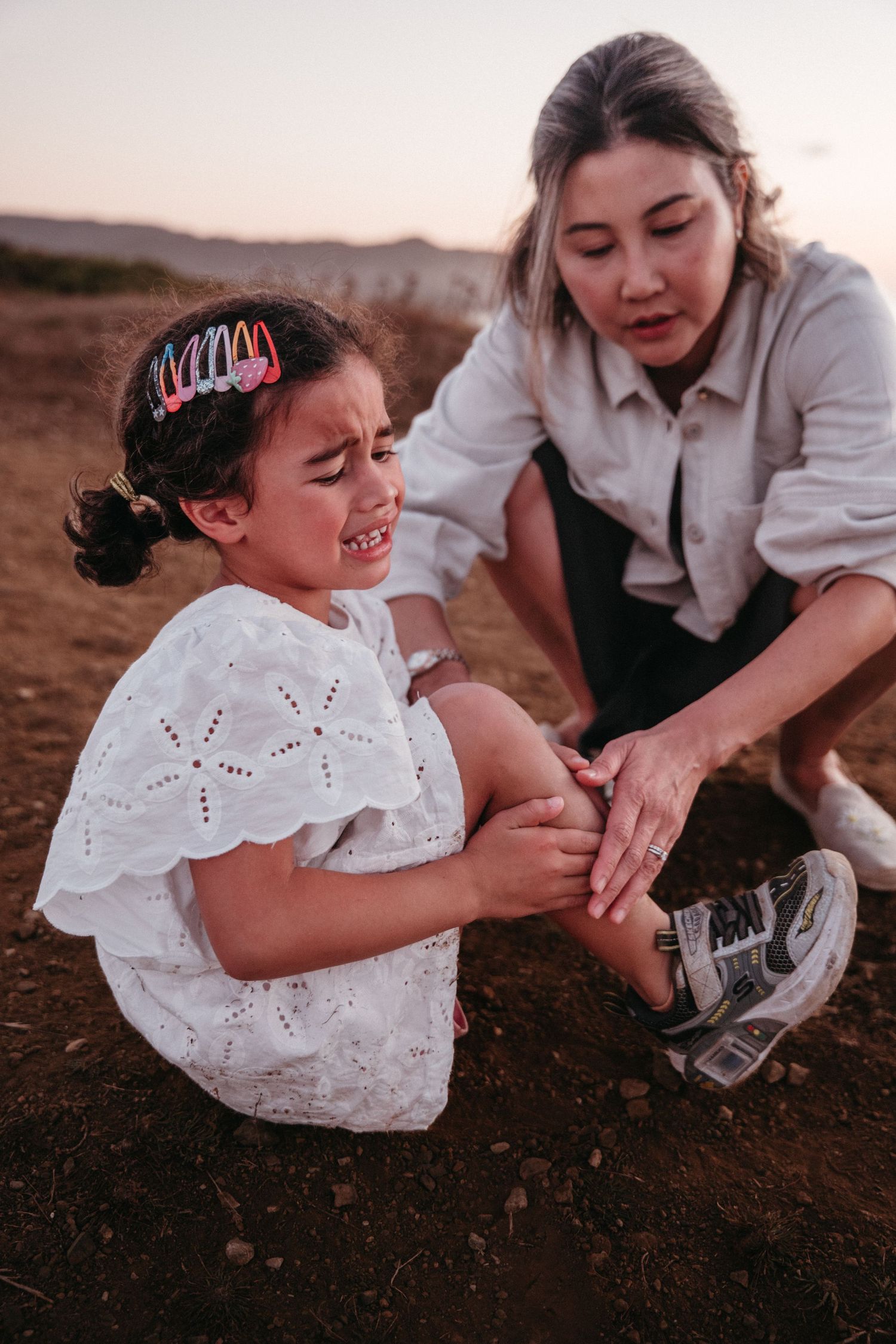 A concerned mother kneels beside her crying young daughter on a dirt path at dusk.