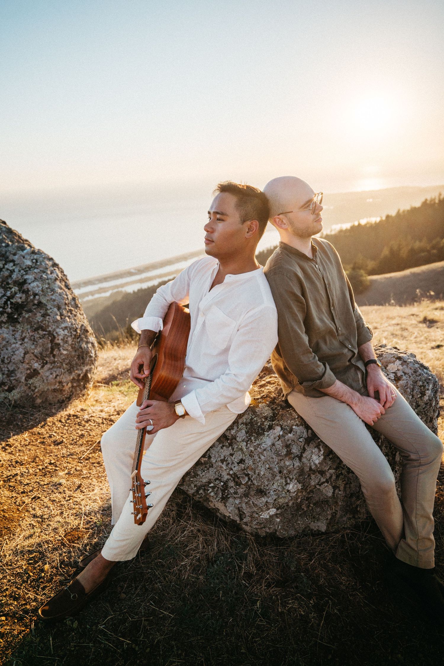 Two musicians sit back-to-back on a rock at sunset, one holding an acoustic guitar, with a scenic coastal view behind them.