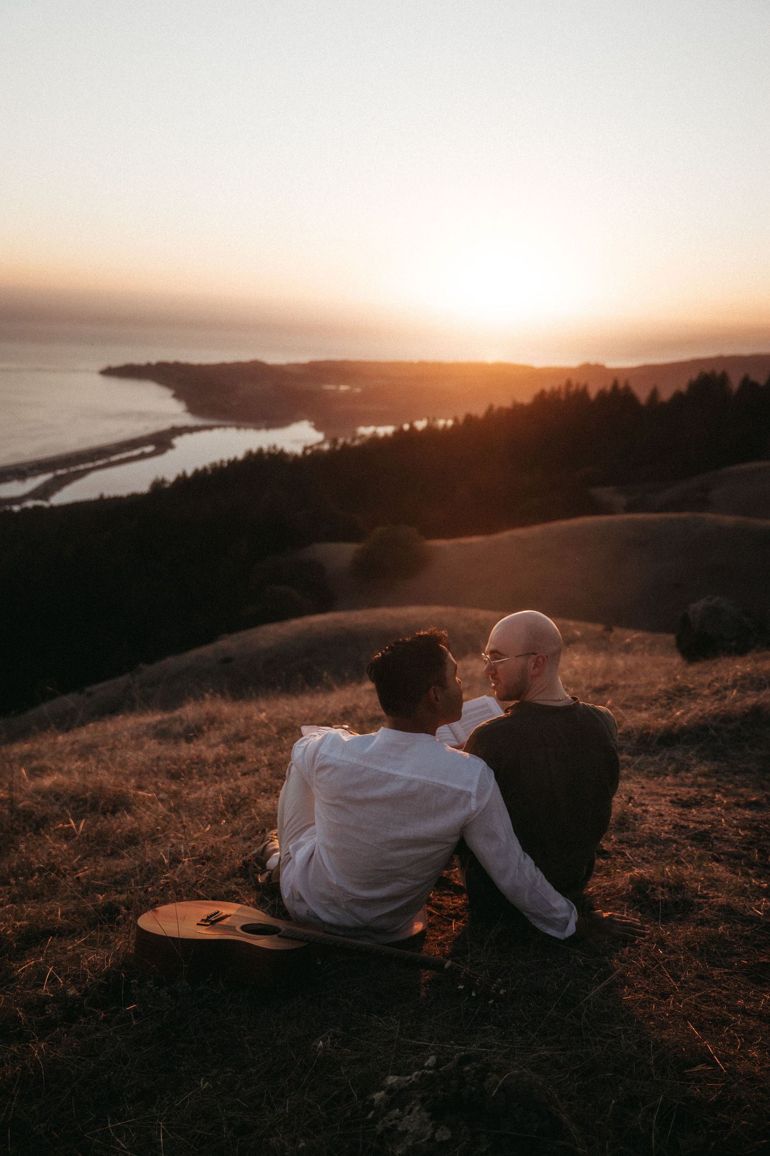 Two people sit on a hillside at sunset, embracing with a guitar beside them and a coastal landscape in the background.
