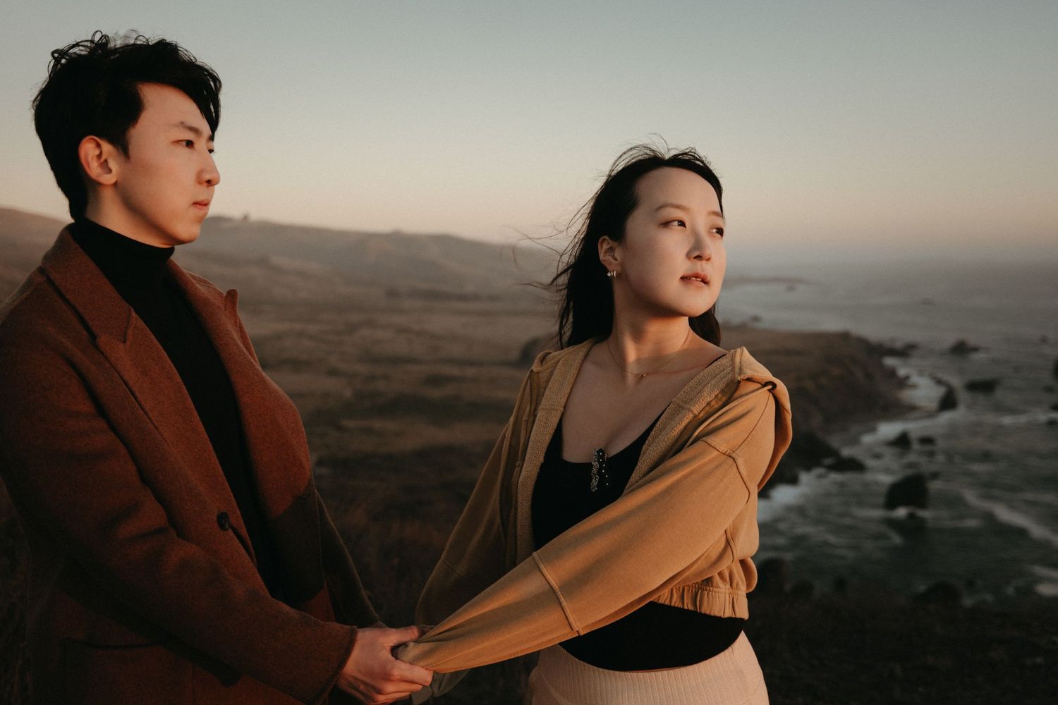 Couple holding hands on a rugged coastal cliff at sunset, one in a rust coat and one in a tan jacket, with the ocean and hills behind.