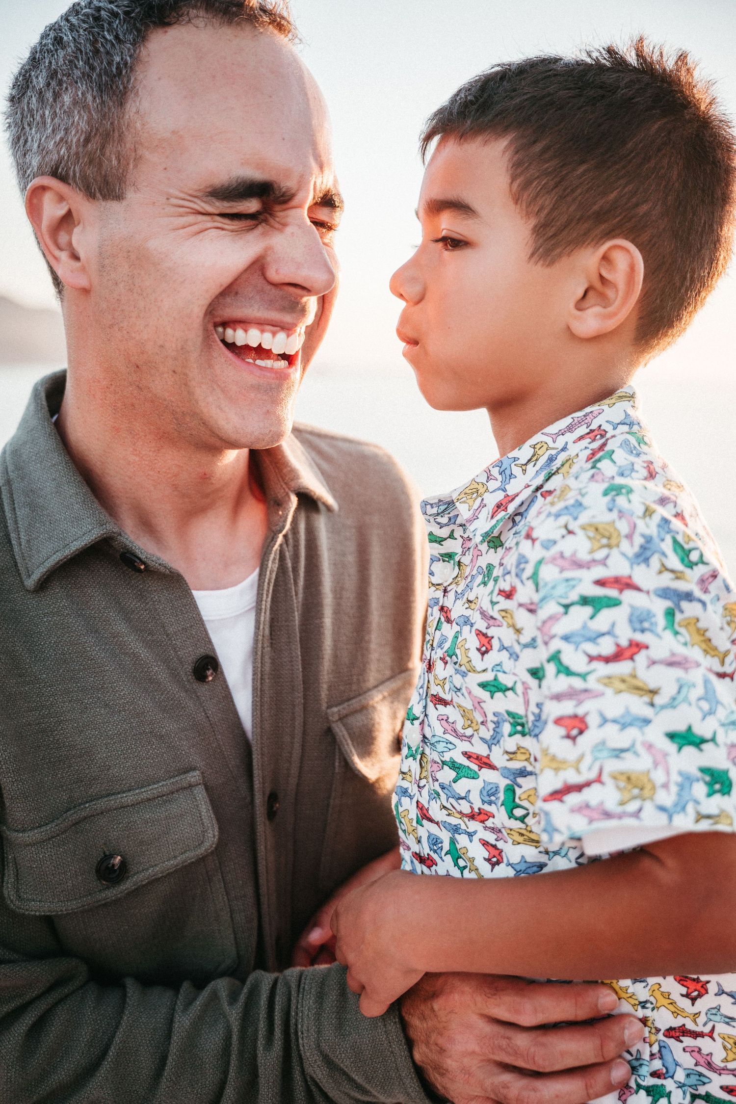 A laughing man holds a young boy in a colorful dinosaur shirt who puckers up for a kiss outdoors.