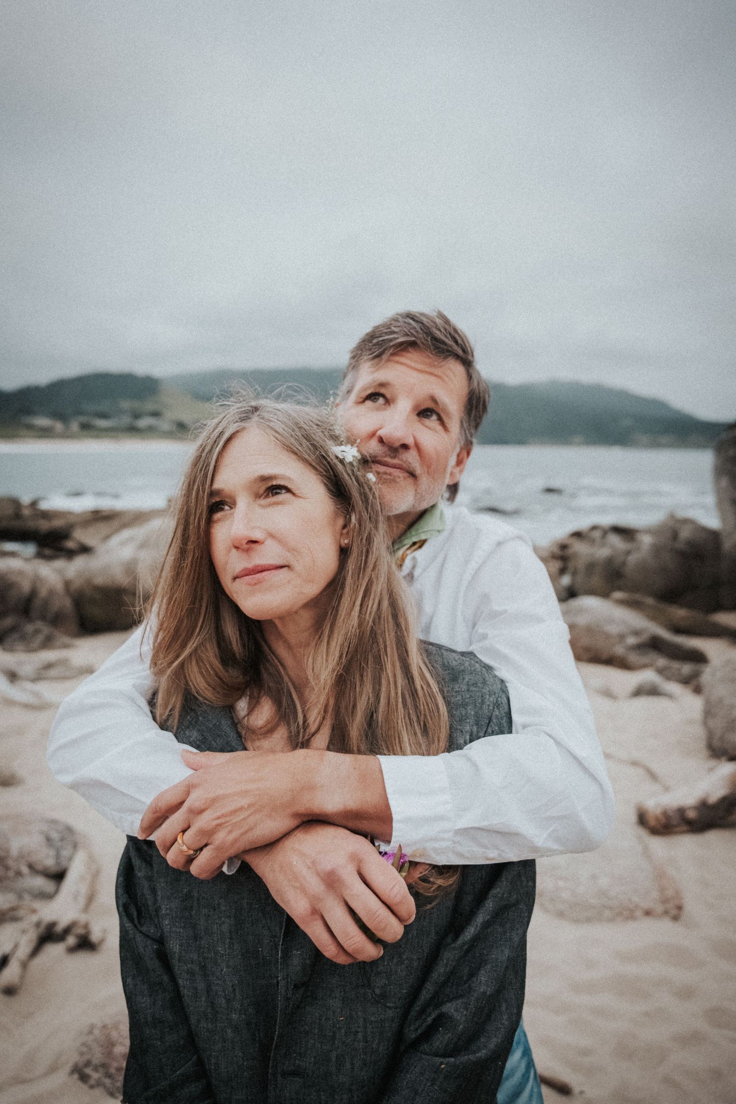 Middle-aged couple embracing on a rocky beach, man in white shirt hugging woman with long hair, overcast coastal scenery.Photography by The
