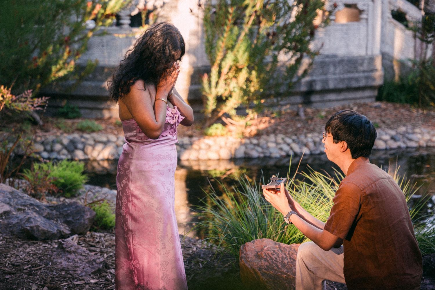 Man kneeling on one knee proposing to woman in pink dress by a garden pond at sunset.