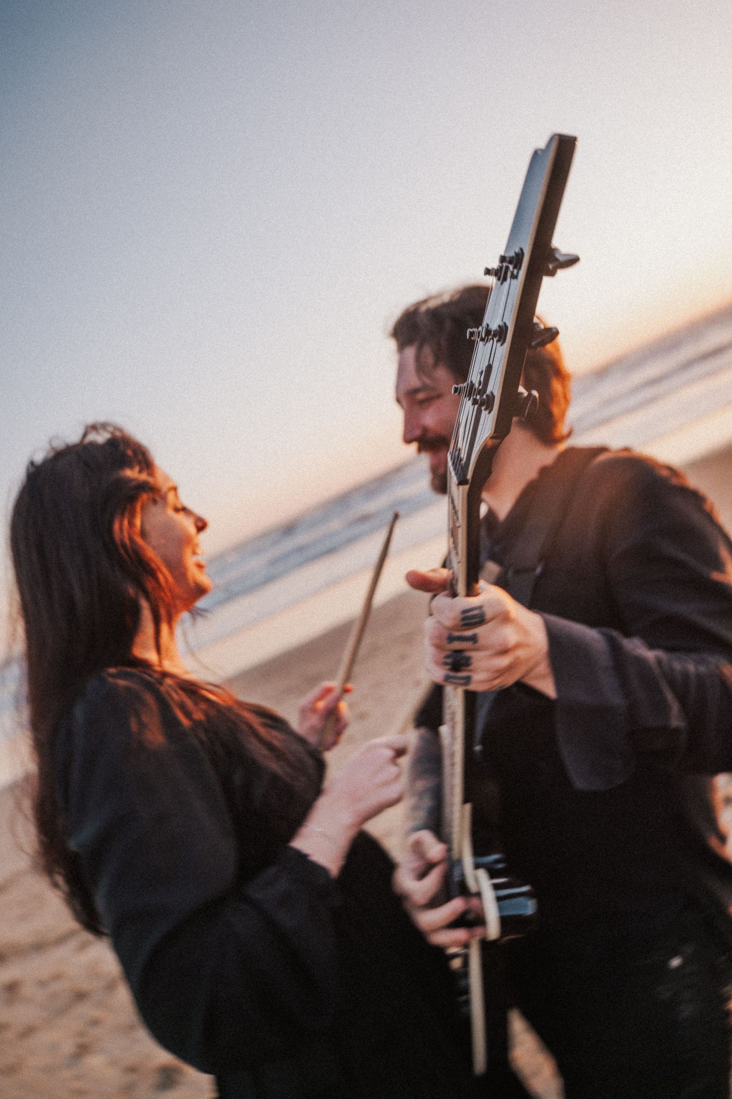 Couple laughing on beach at sunset, man holding black electric guitar, woman with drumsticks, ocean in background. Photography by The Y.K. S