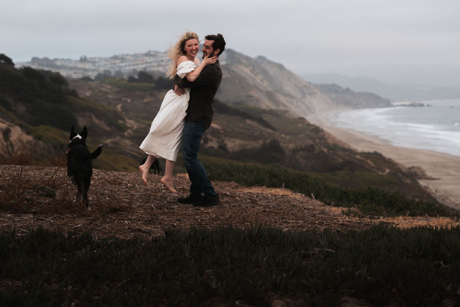 Couple dancing on coastal cliffside with dog, ocean backdrop, romantic outdoor engagement photo.