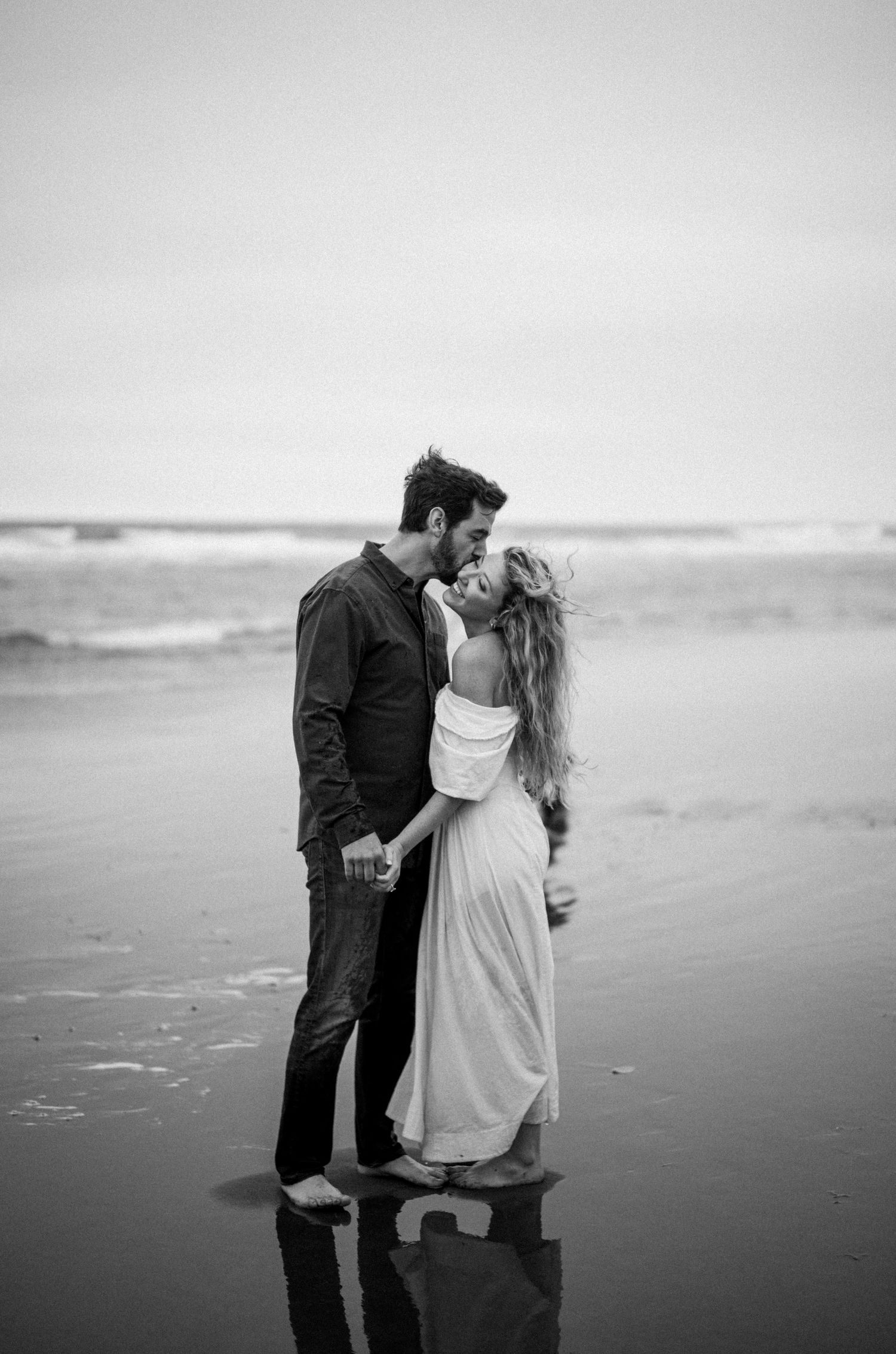 Black and white romantic photo of a couple kissing on a reflective wet beach at the shoreline.