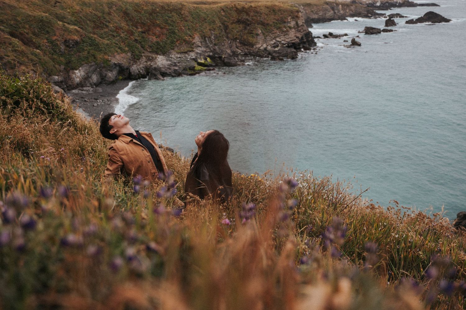 Couple sitting on a scenic coastal hillside with wildflowers, overlooking turquoise ocean waters and rocky shoreline.