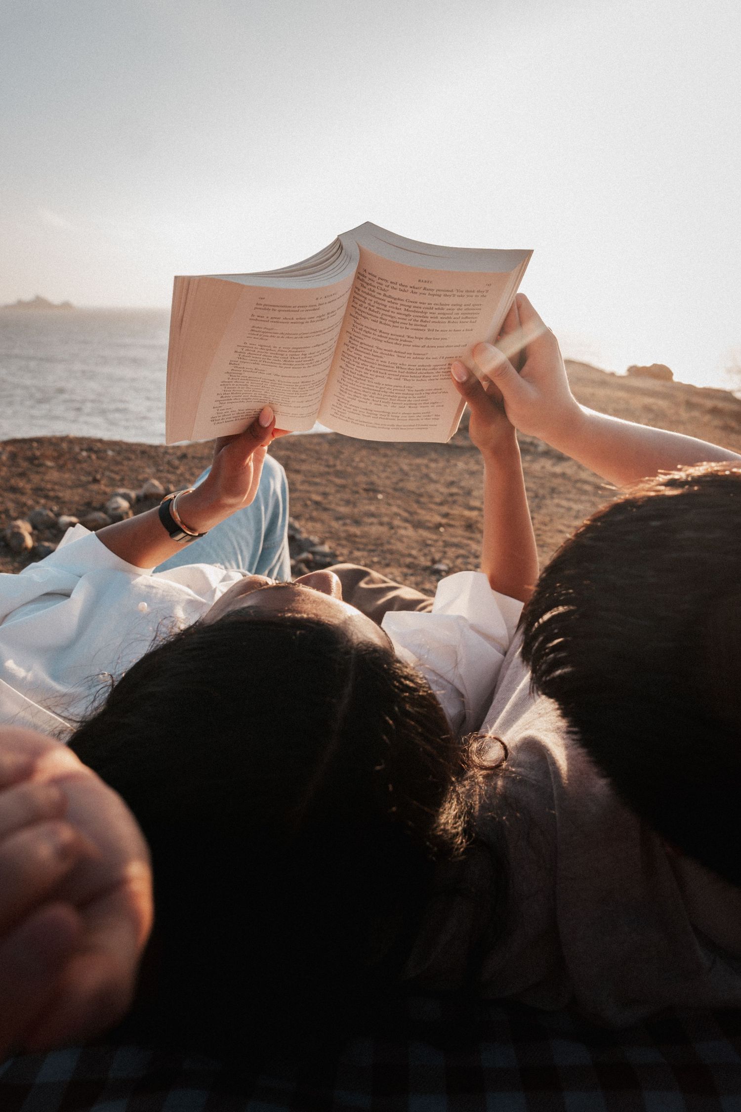 Two people lying outdoors by the sea, sharing a book together in warm golden sunlight.