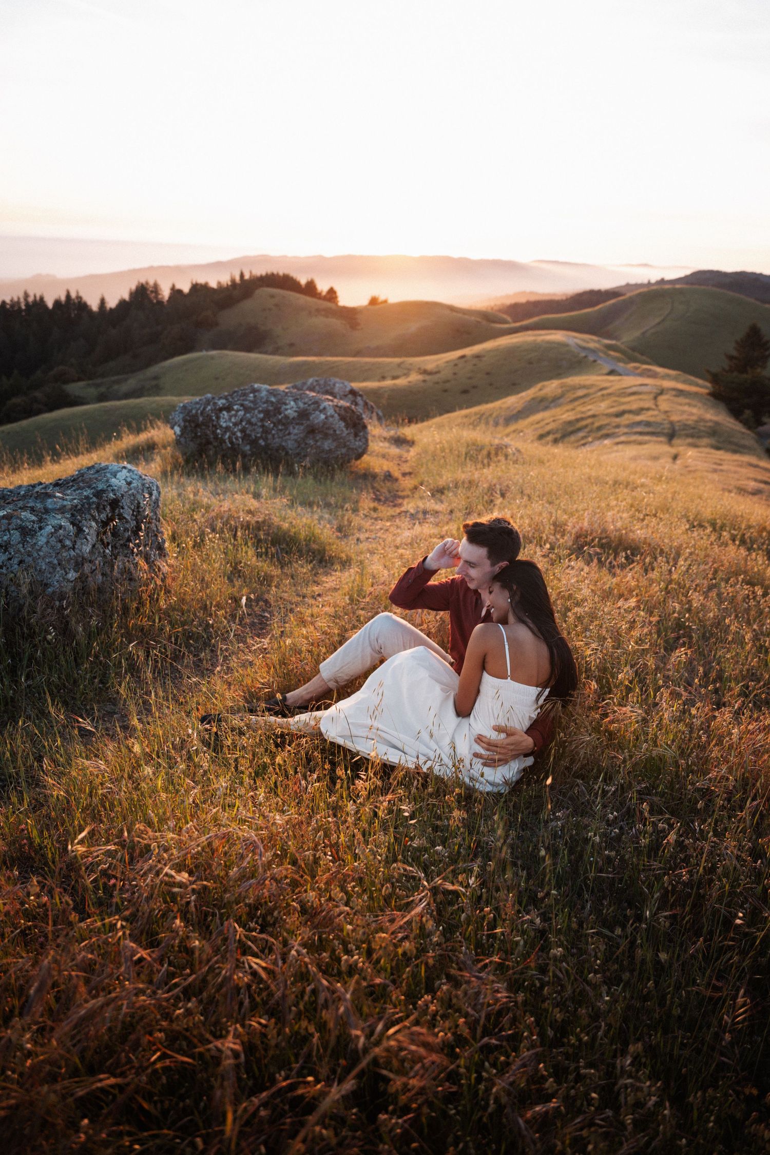 Couple sitting in golden grass field at sunset, embracing romantically with rolling green hills and warm light in background.