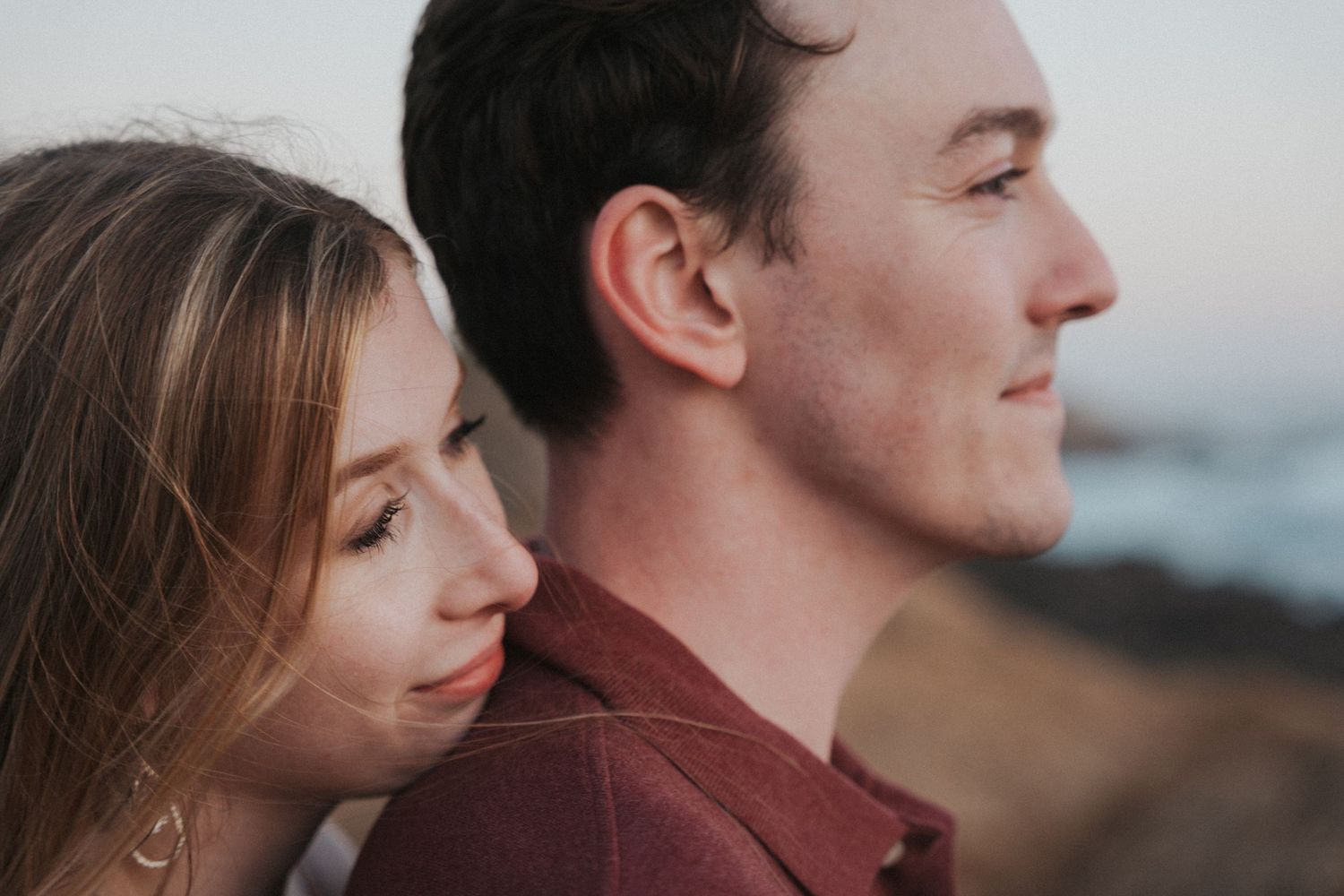 A couple shares a tender moment outdoors at sunset, woman resting her head near man's shoulder.
