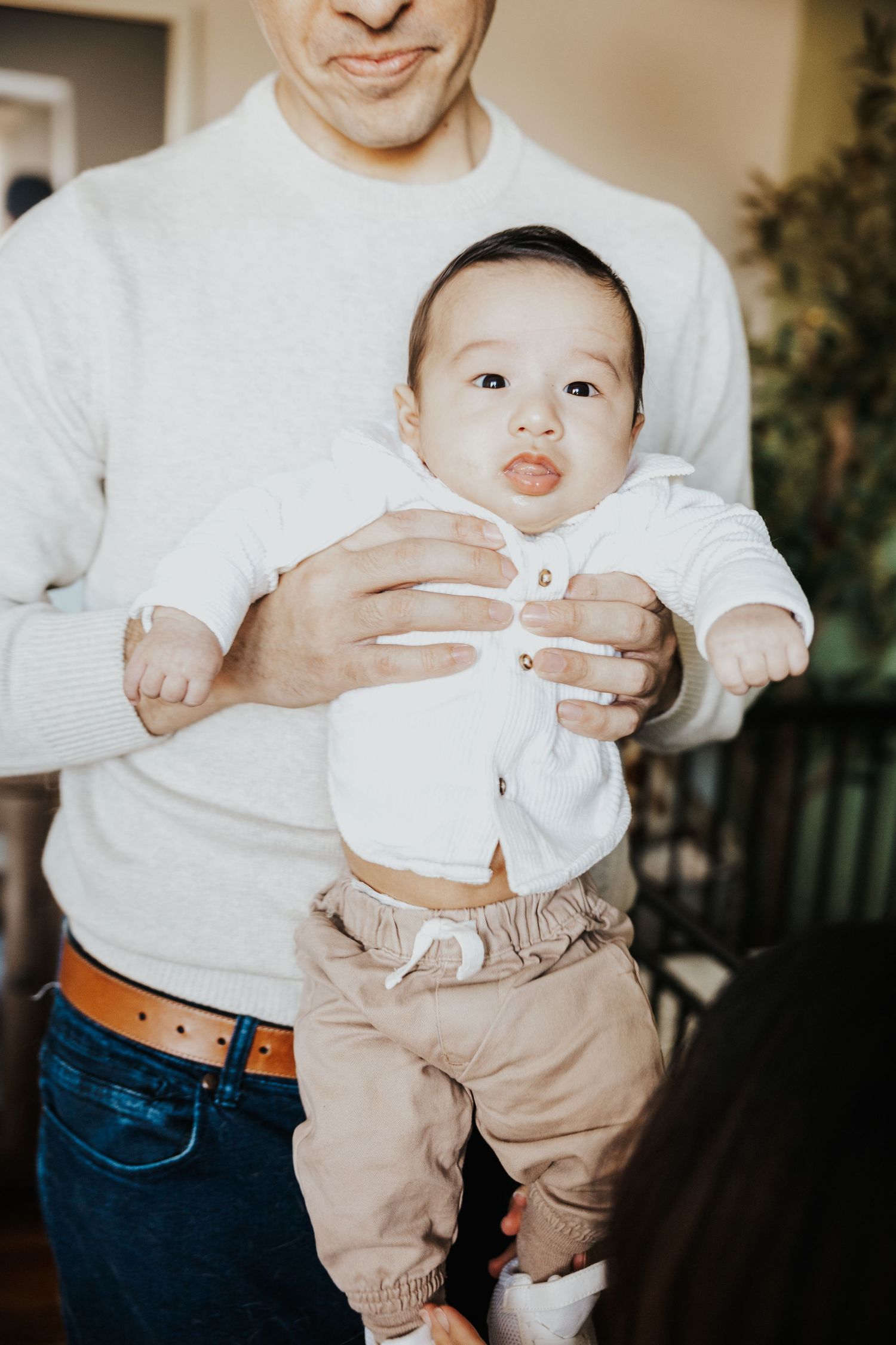 Father in white sweater holding adorable baby in white cardigan and tan pants, smiling indoors.