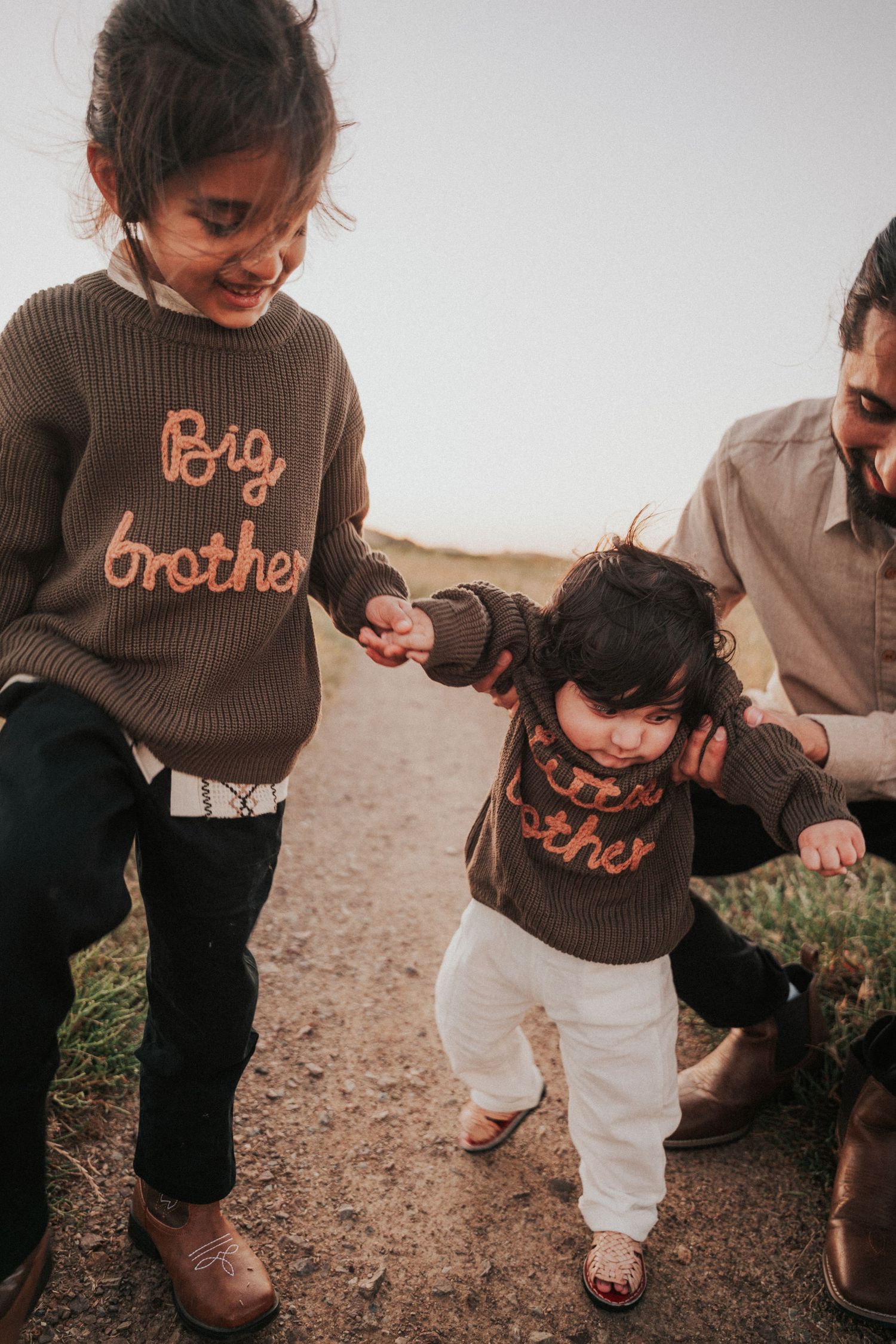 Two children in matching Big Brother and Little Brother sweaters hold hands on a dirt path with an adult nearby.