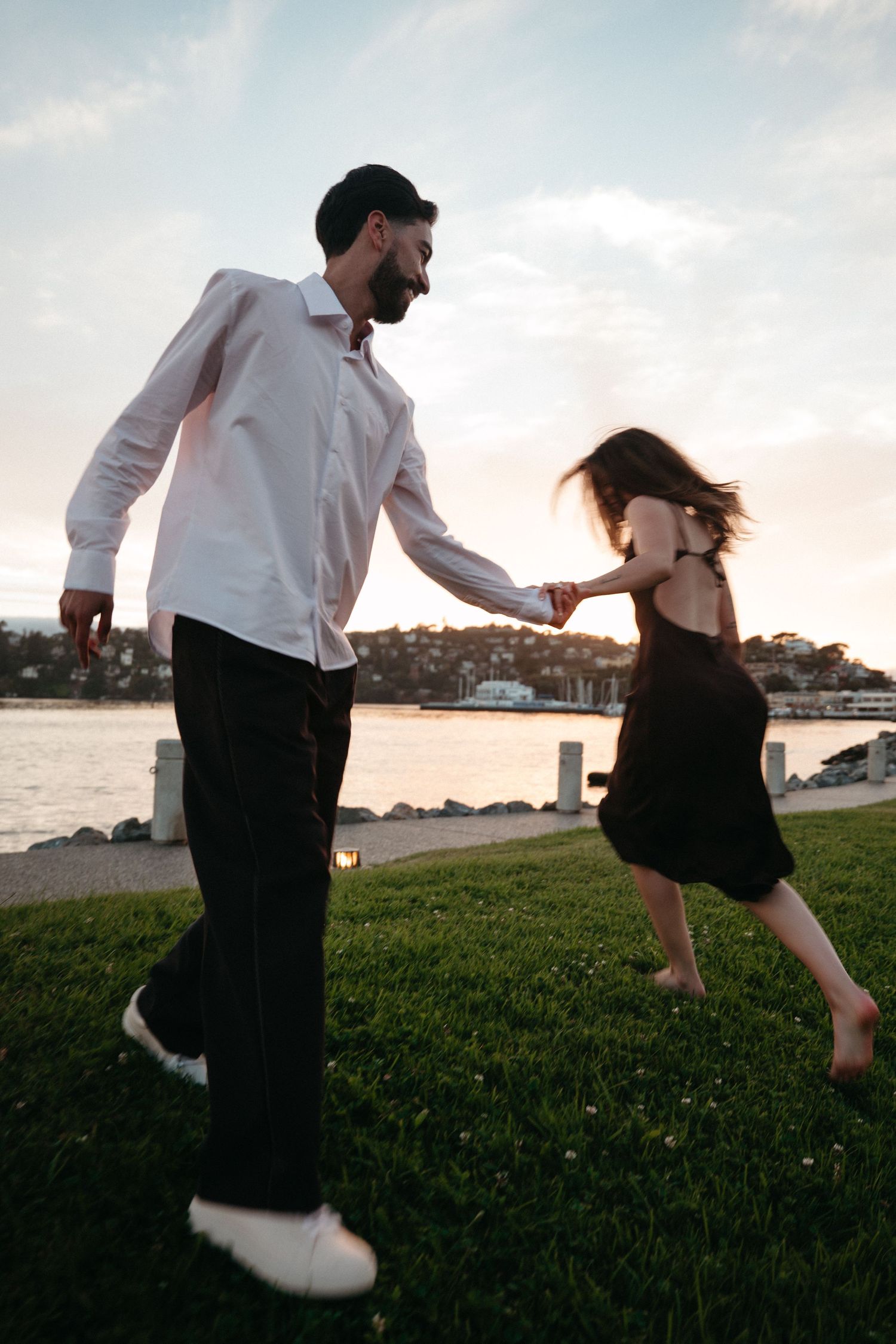 A couple dances joyfully on a grassy waterfront at sunset, man in white shirt and woman in black dress holding hands.