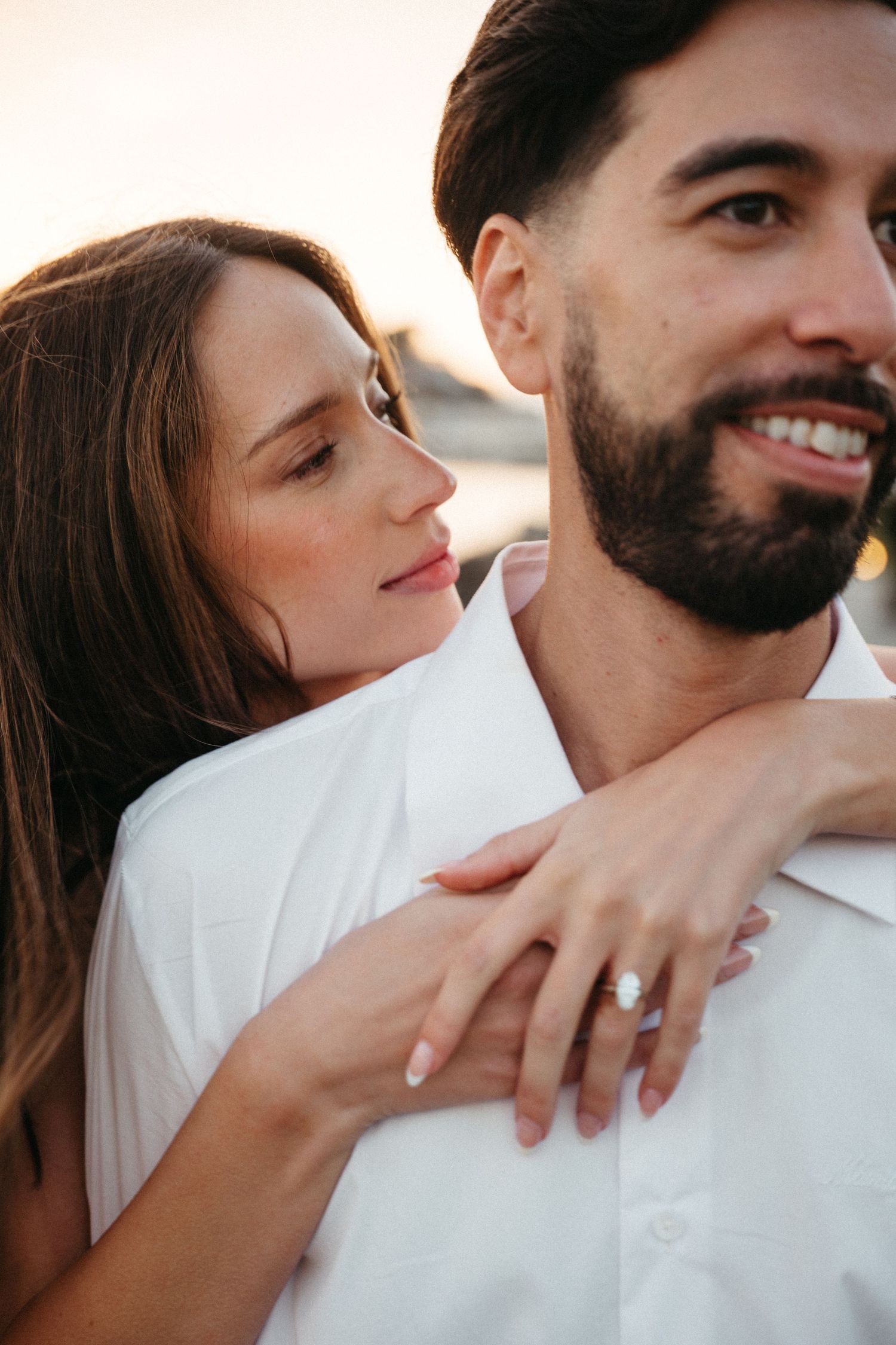 Couple in white embracing at sunset, woman showing diamond engagement ring while man smiles warmly.