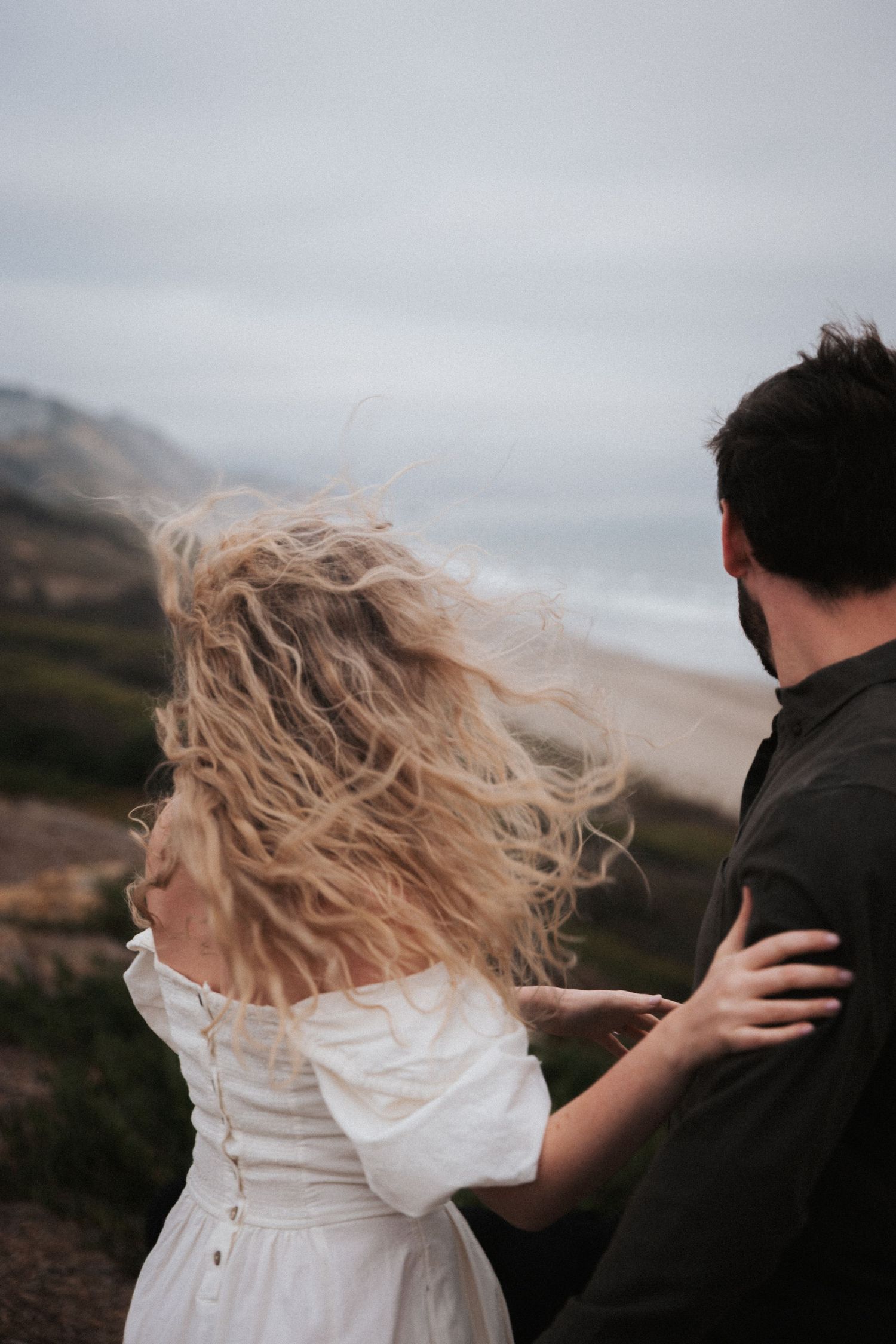 Couple embracing on a windy coastal cliff, woman in white dress with blonde hair blowing, ocean backdrop.