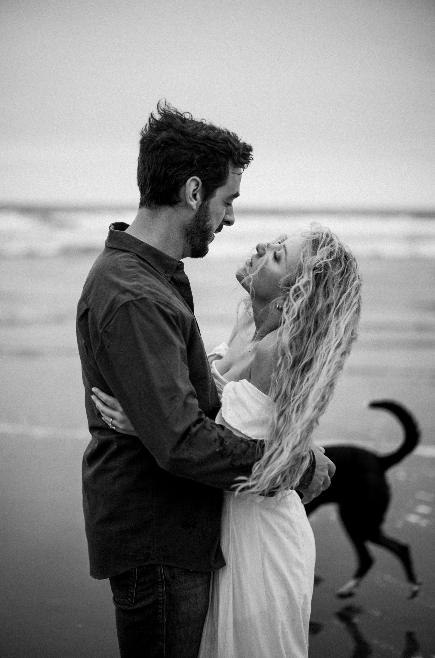Black and white photo of couple embracing and laughing on beach with a dog in the background near ocean waves.