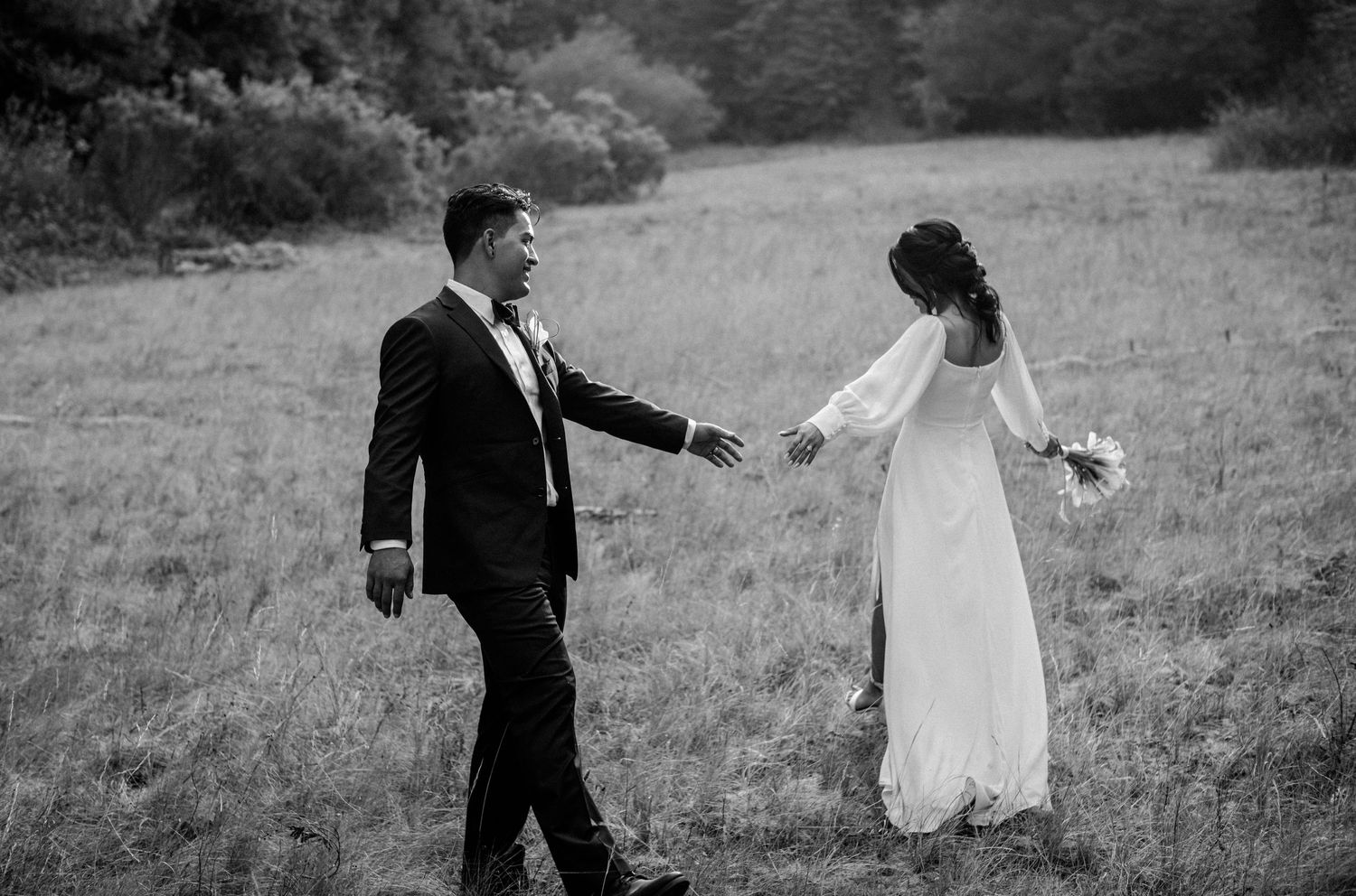 Black and white photo of a bride and groom walking hand in hand through a lush green field on their wedding day.