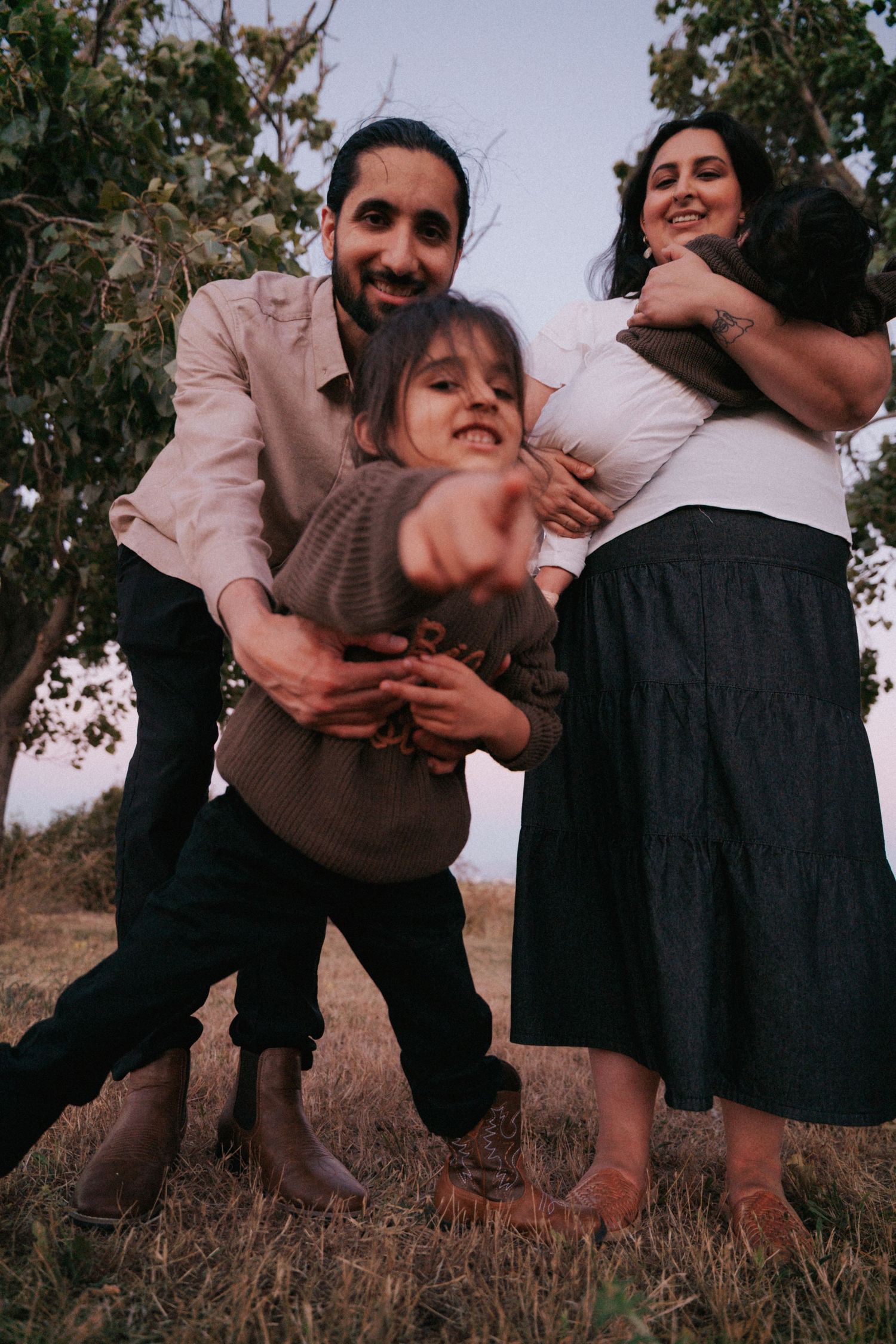 Joyful family of four laughing and embracing outdoors in a grassy field with trees in background.