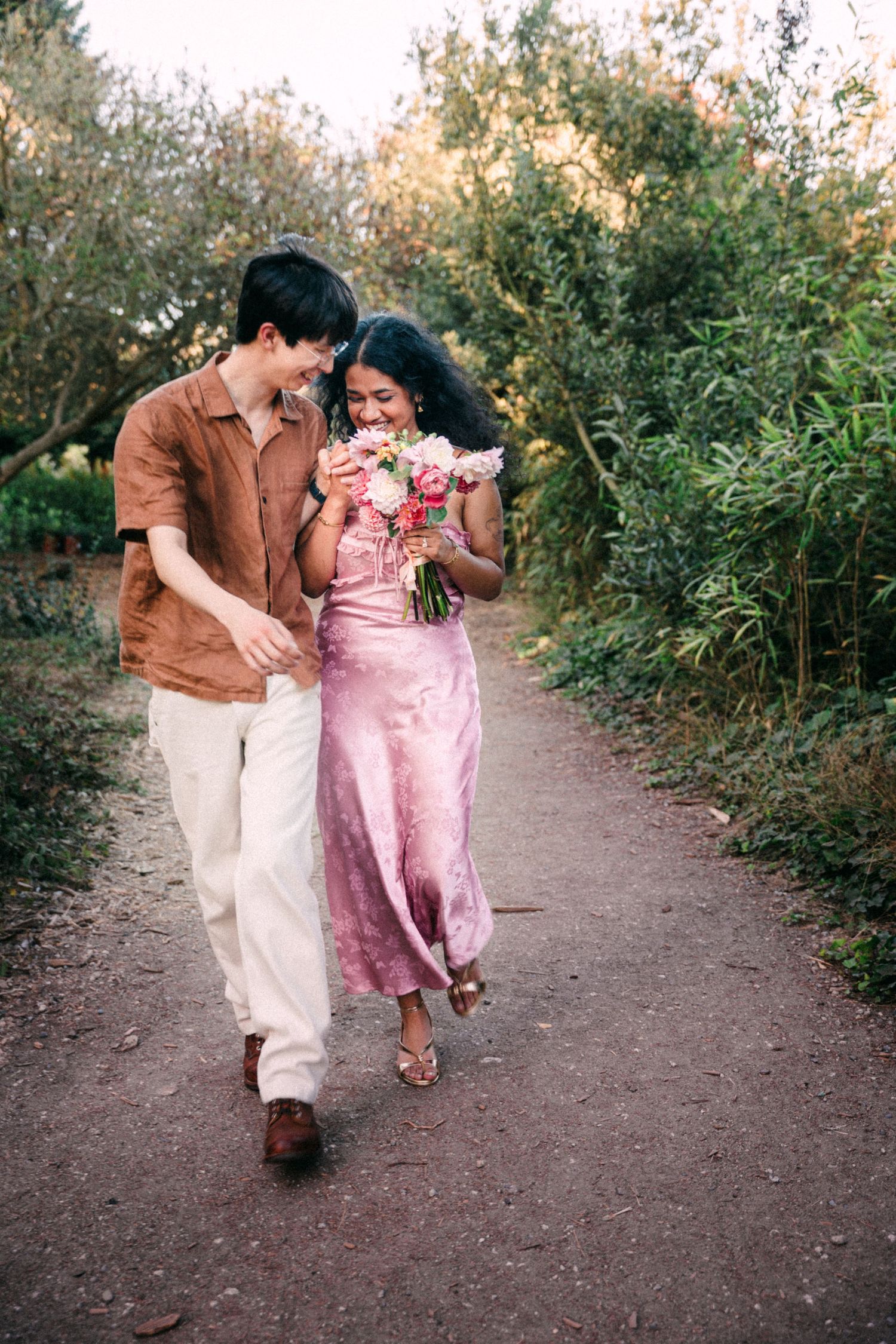Couple walking on garden path, woman in pink dress holding floral bouquet, man in brown shirt and white pants.