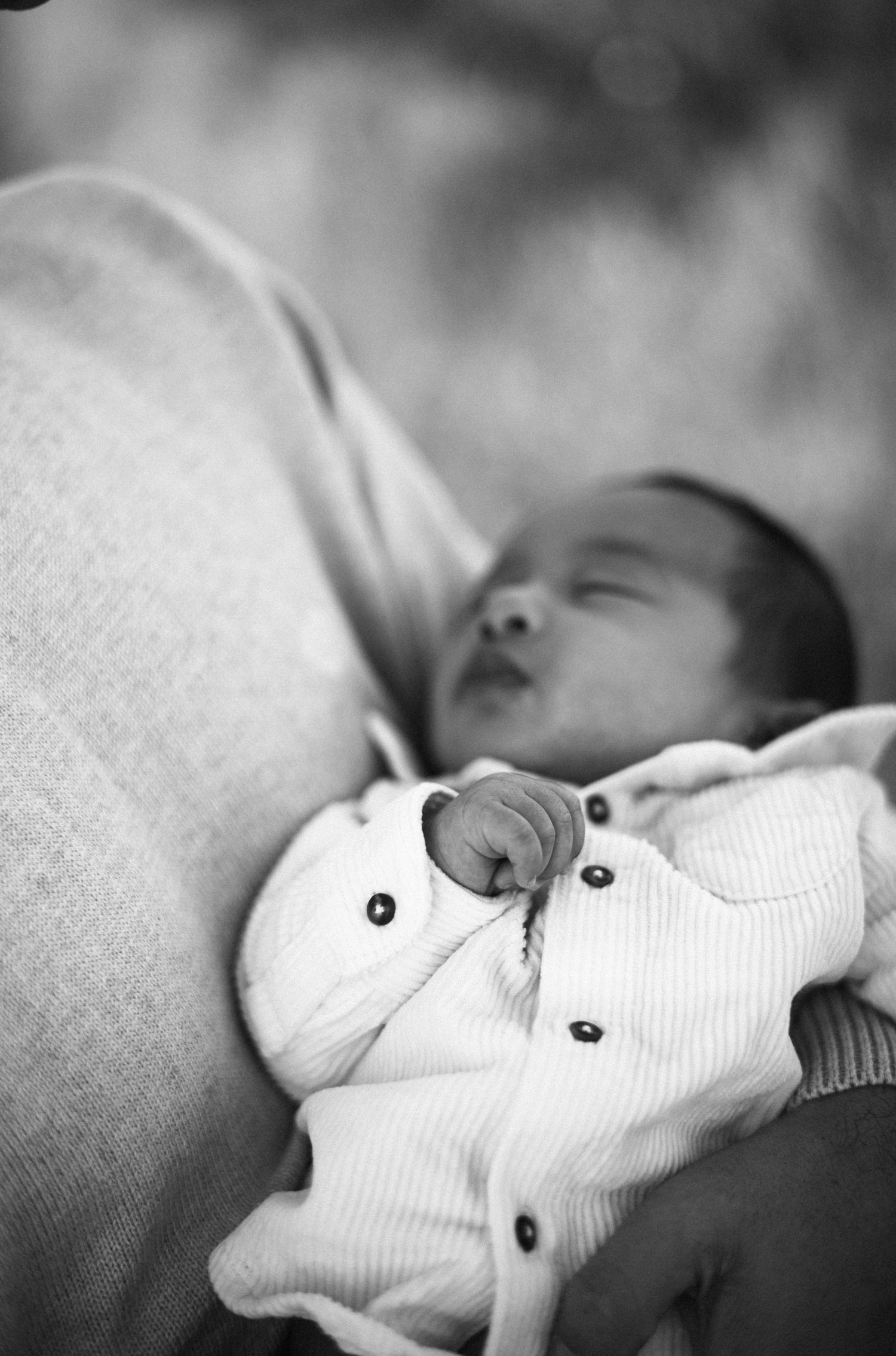 Sleeping newborn baby cradled in parent's arm wearing white outfit in black and white photo.