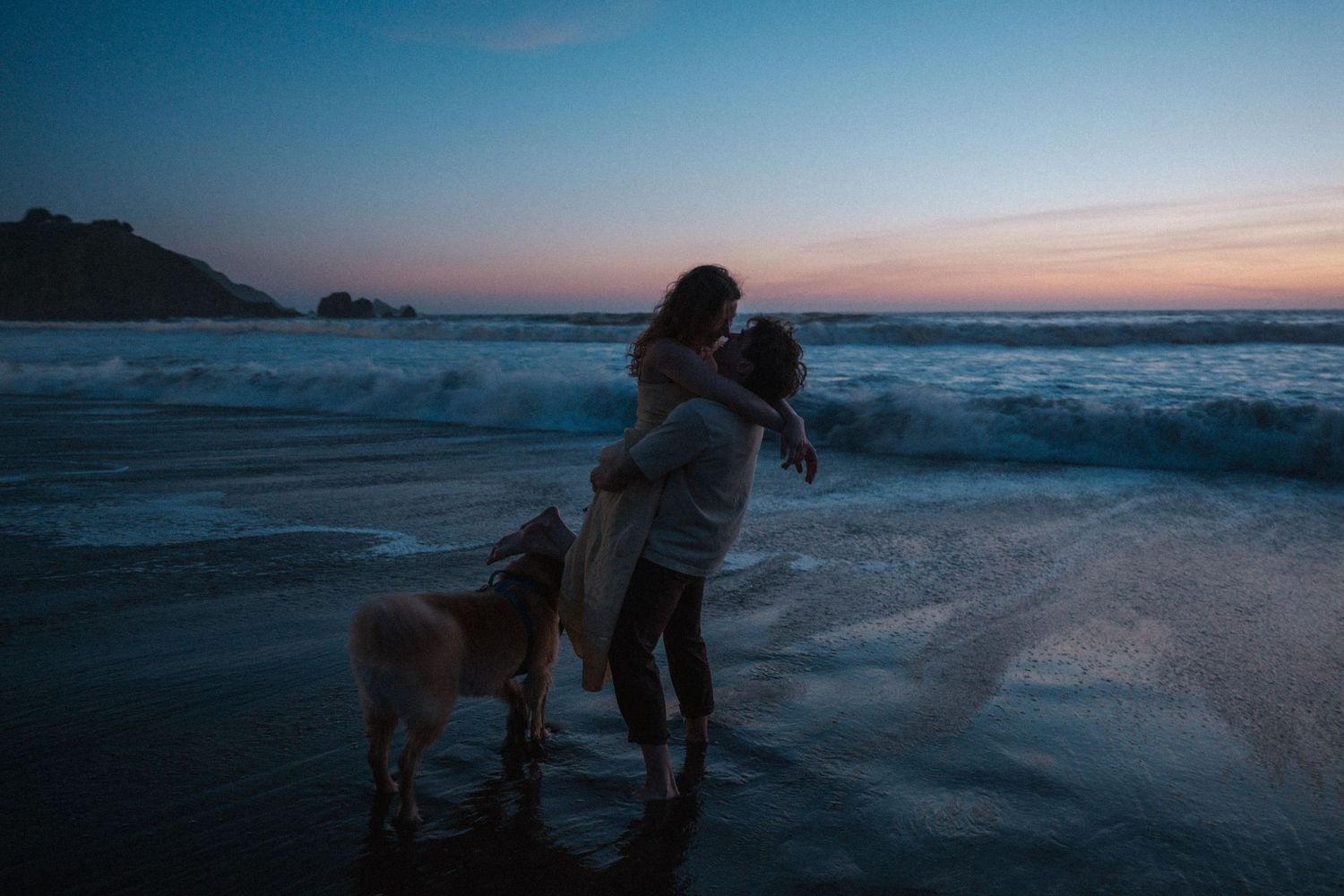 Romantic couple embracing at ocean shore during sunset with golden retriever beside them.