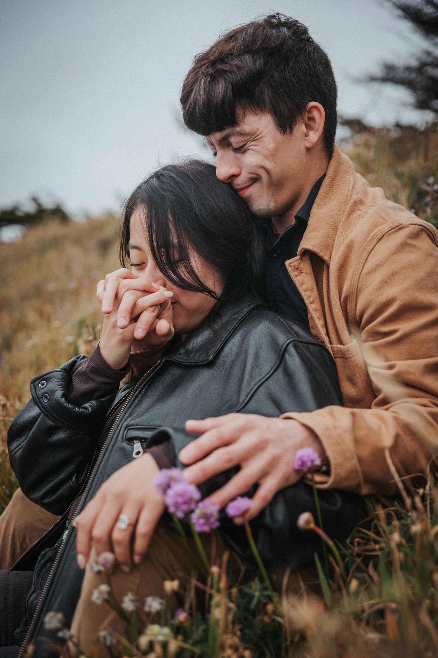 Couple sitting in a field of wildflowers, embracing tenderly as man nuzzles woman in leather jacket.